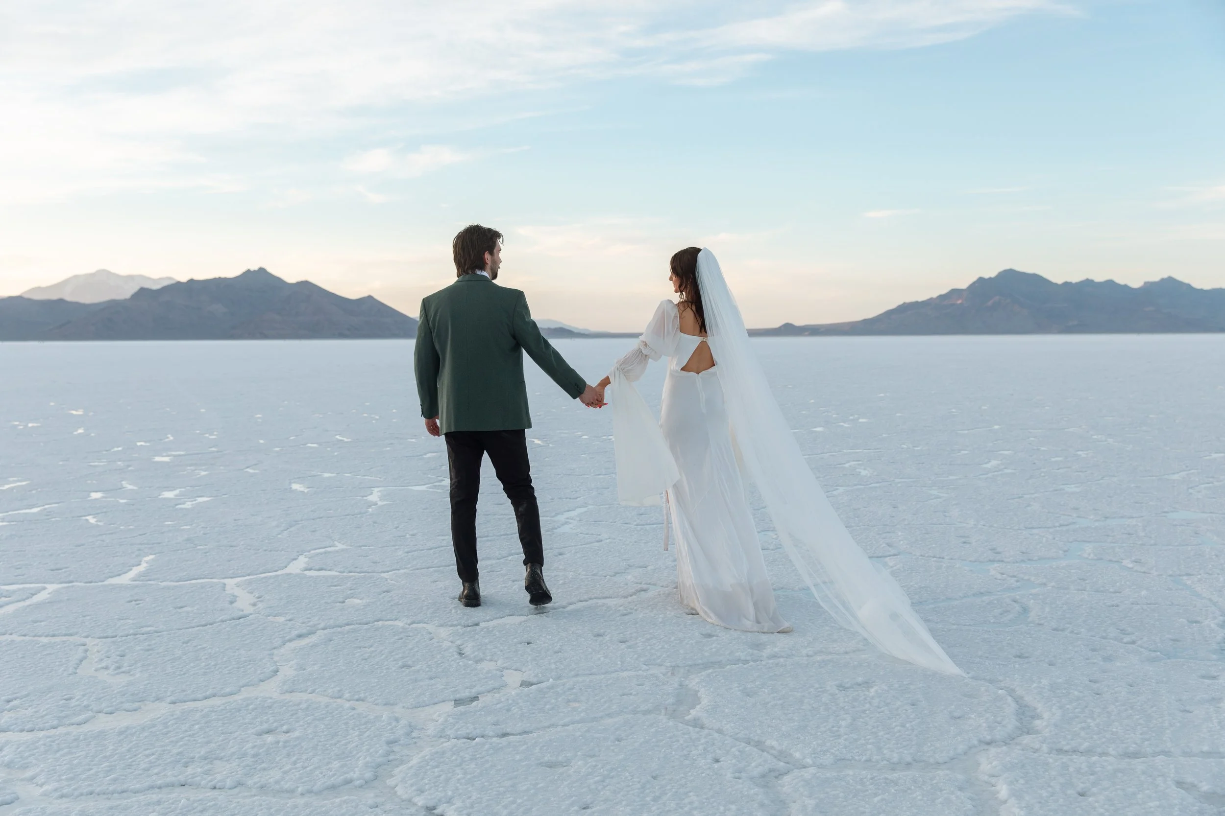 A couple dressed in wedding attire holding hands and walking on a salt flat with mountains in the background.
