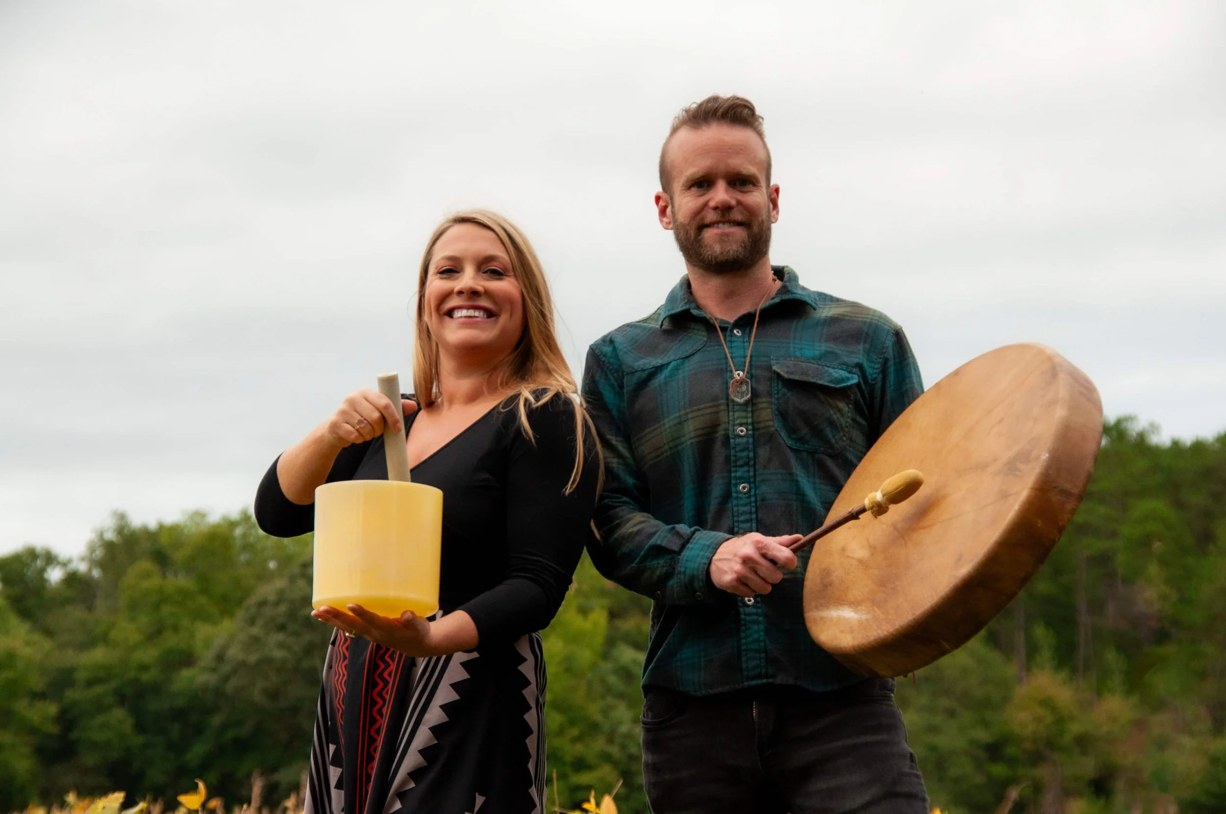 A woman and a man standing outdoors overcast sky, holding musical instruments and smiling at the camera. The woman holds a yellow glass bowl with a wooden mallet, and the man holds a large wooden drum and a wooden drumstick.