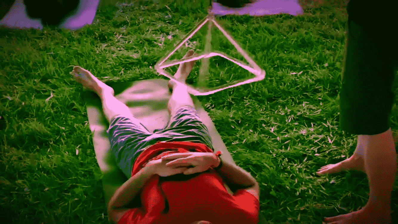 A person lying on the grass in front of a glowing geometric star-shaped object, with another person standing nearby.