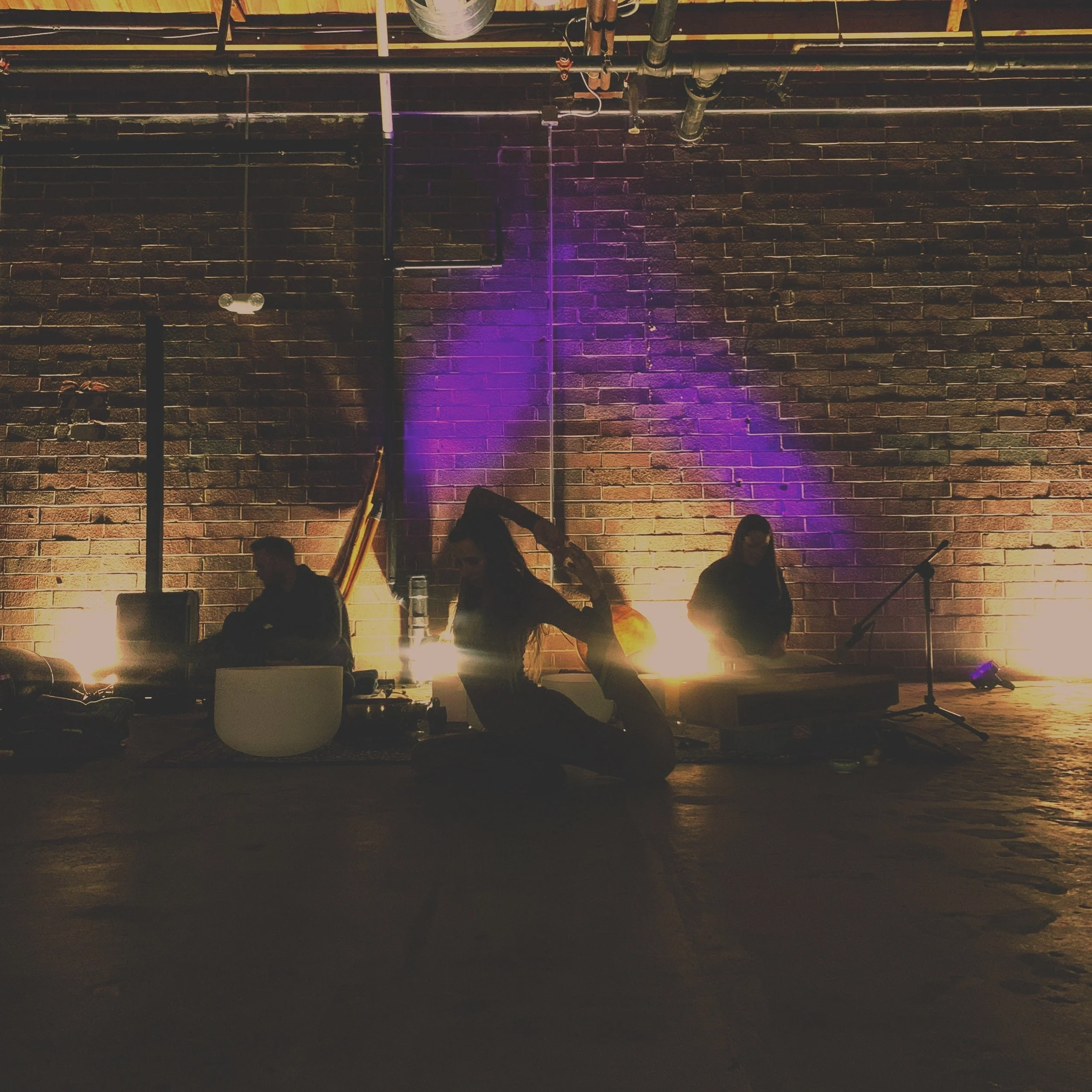 Three musicians playing instruments in front of a brick wall illuminated by purple and yellow lighting, with a person seated on the floor adjusting hair.