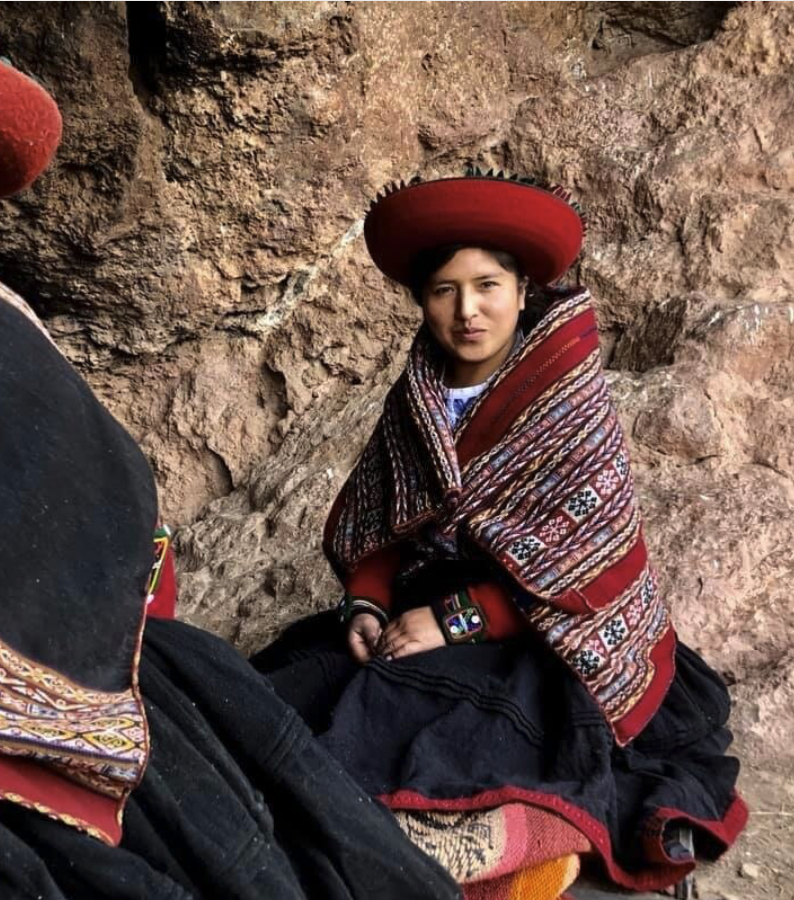 A young woman wearing traditional Andean clothing, including a large red hat and a colorful woven shawl, sitting against a rocky background.