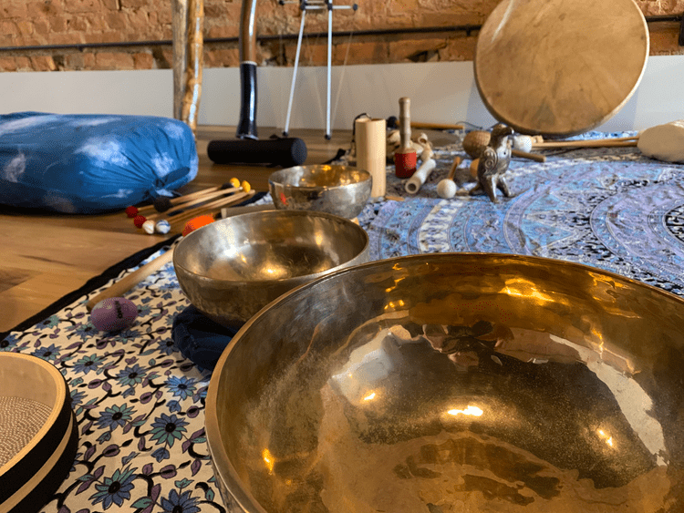 Multiple singing bowls of various sizes on a patterned cloth, with percussion mallets and other objects scattered in a room with a brick wall background.