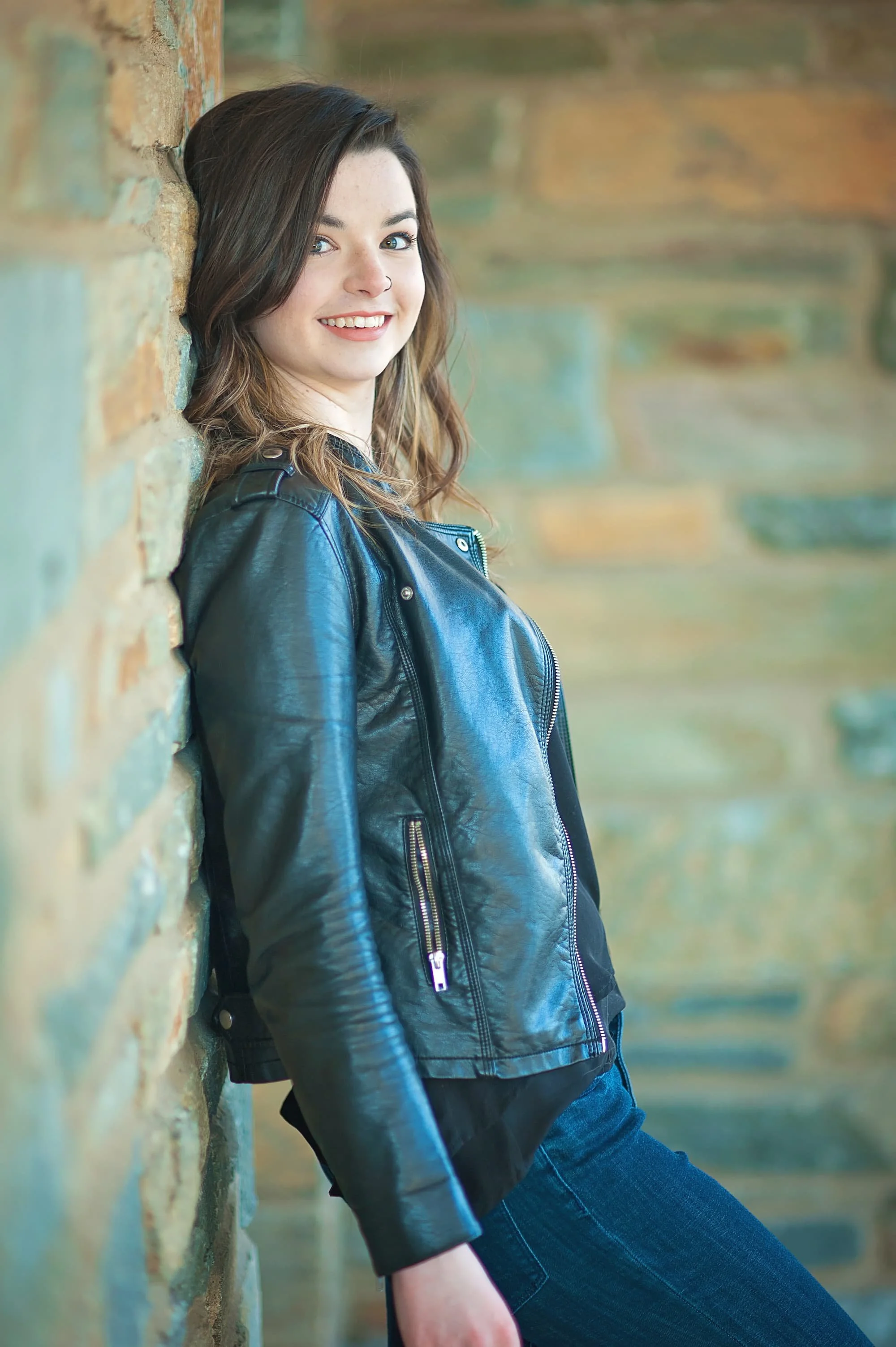 Senior portrait of a girl wearing a black leather jacket leaning against a stone wall