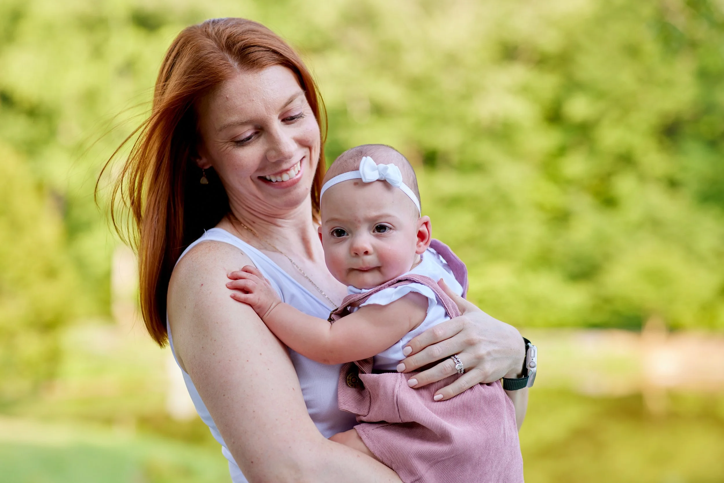 Smiling woman holding a baby with a headband in an outdoor setting.