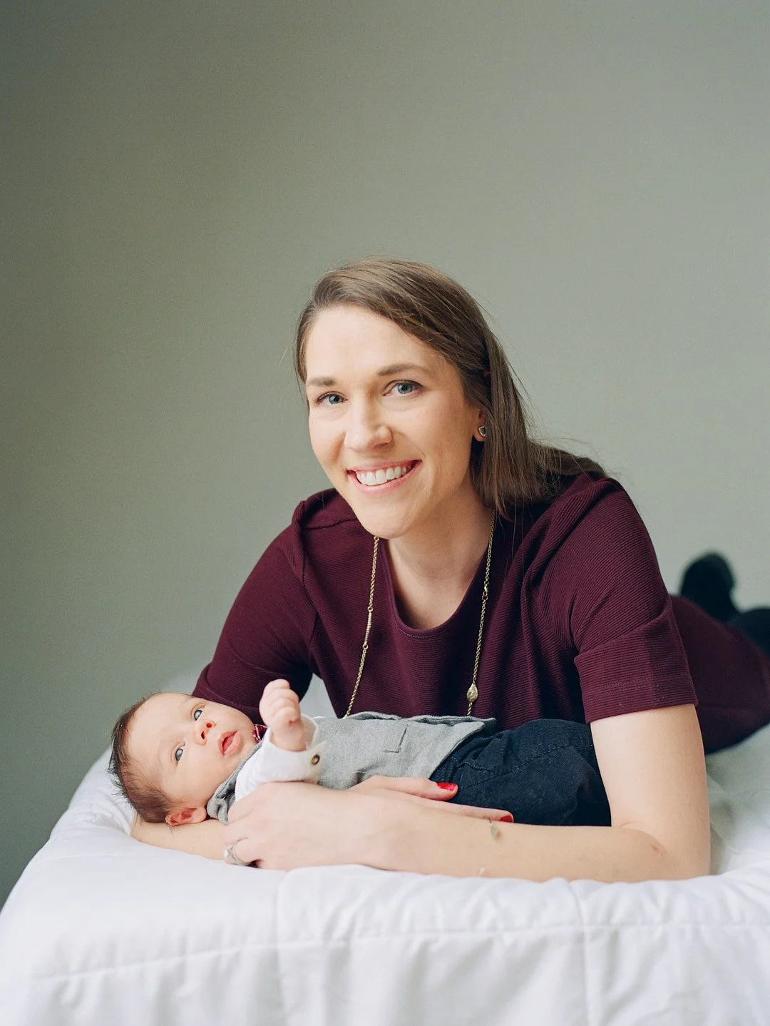 Portrait of a mom wearing a burgundy dress laying on a bed with her newborn baby