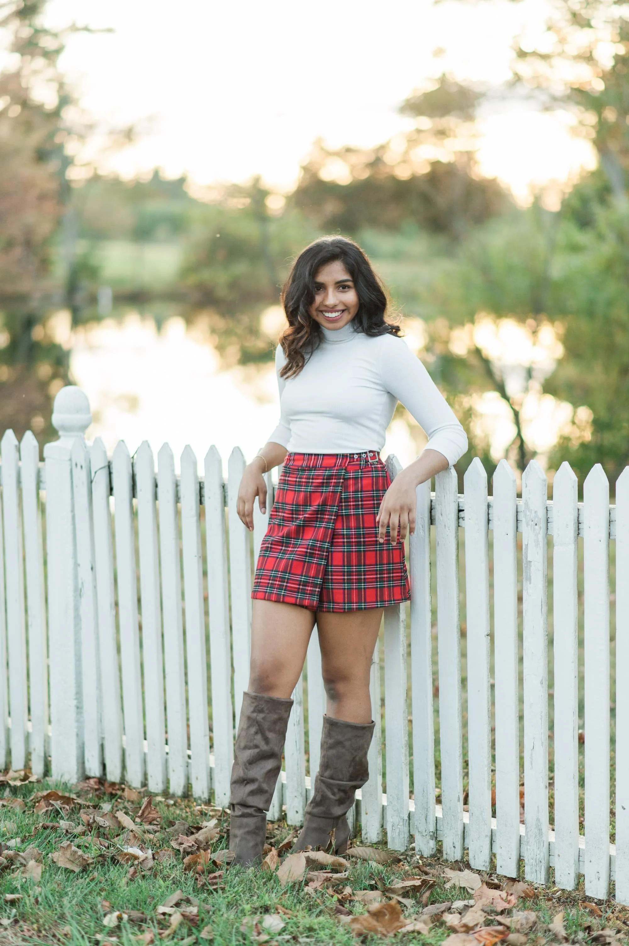 Senior portraits of a girl wearing a plaid skirt leaning against a white fence