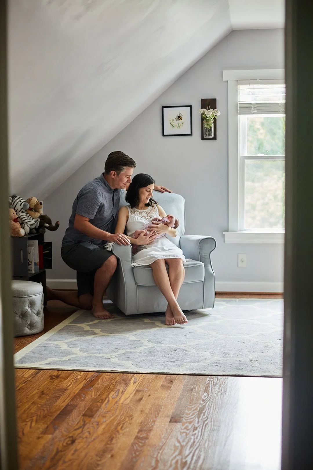 Portrait of mom, dad and baby in the nursery. Mom is sitting in a rocking chair near a window with dad leaning over the arm.