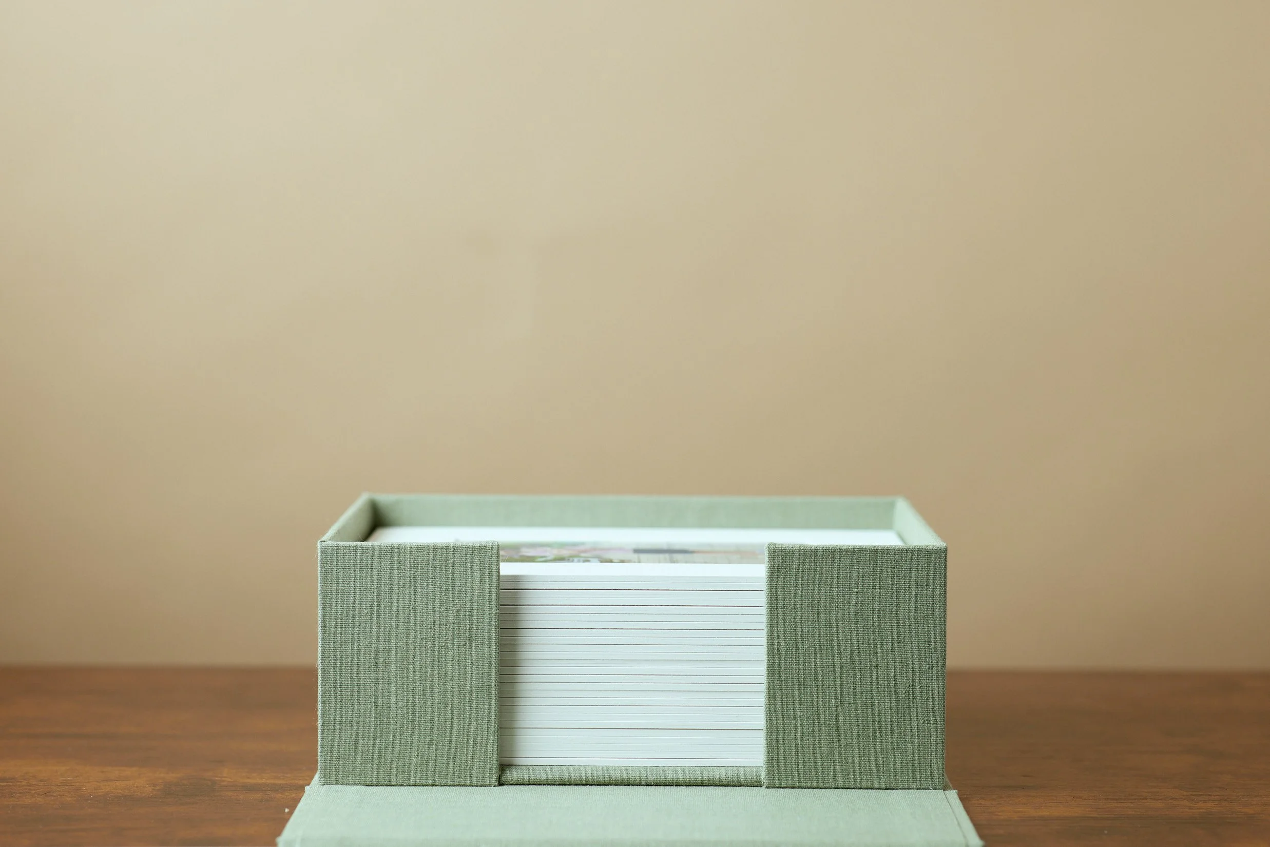 A green fabric-covered folio box holding a stack of prints with colorful designs on top, placed on a wooden surface against a plain beige wall.