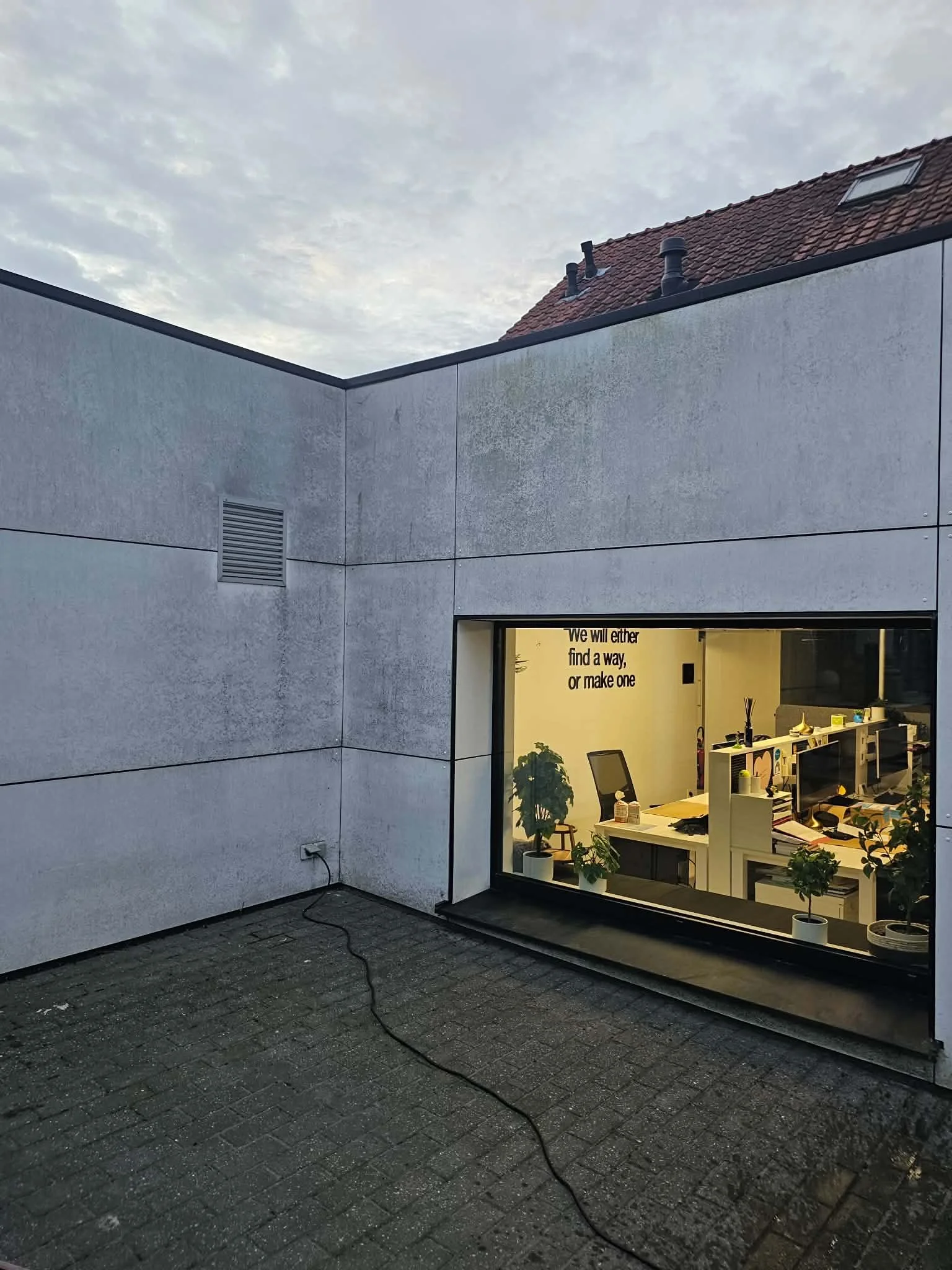 Uitzicht op de buitenkant van een modern kantoor met grote ramen, te zien vanuit een donker stenen patio, met een lucht vol wolken erboven.