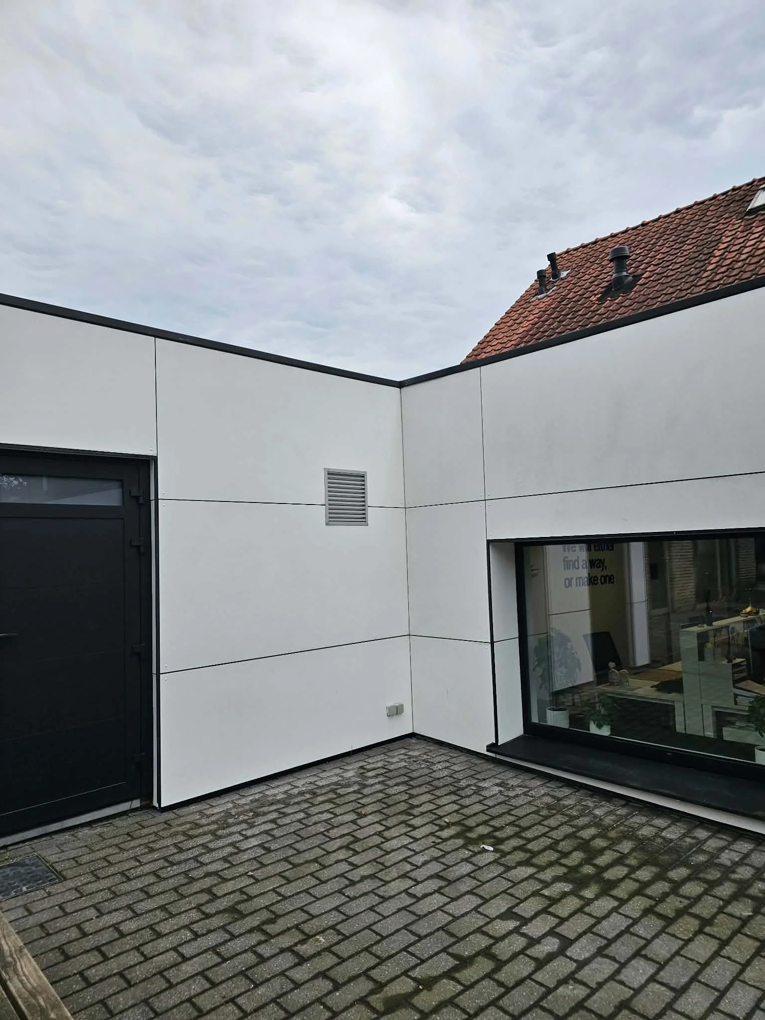 Moderne witte bouw met grote ramen, een zwart schuifdeur, een ventilatierooster en een rieten dak met twee schoorstenen, gelegen op een stenen oprit.