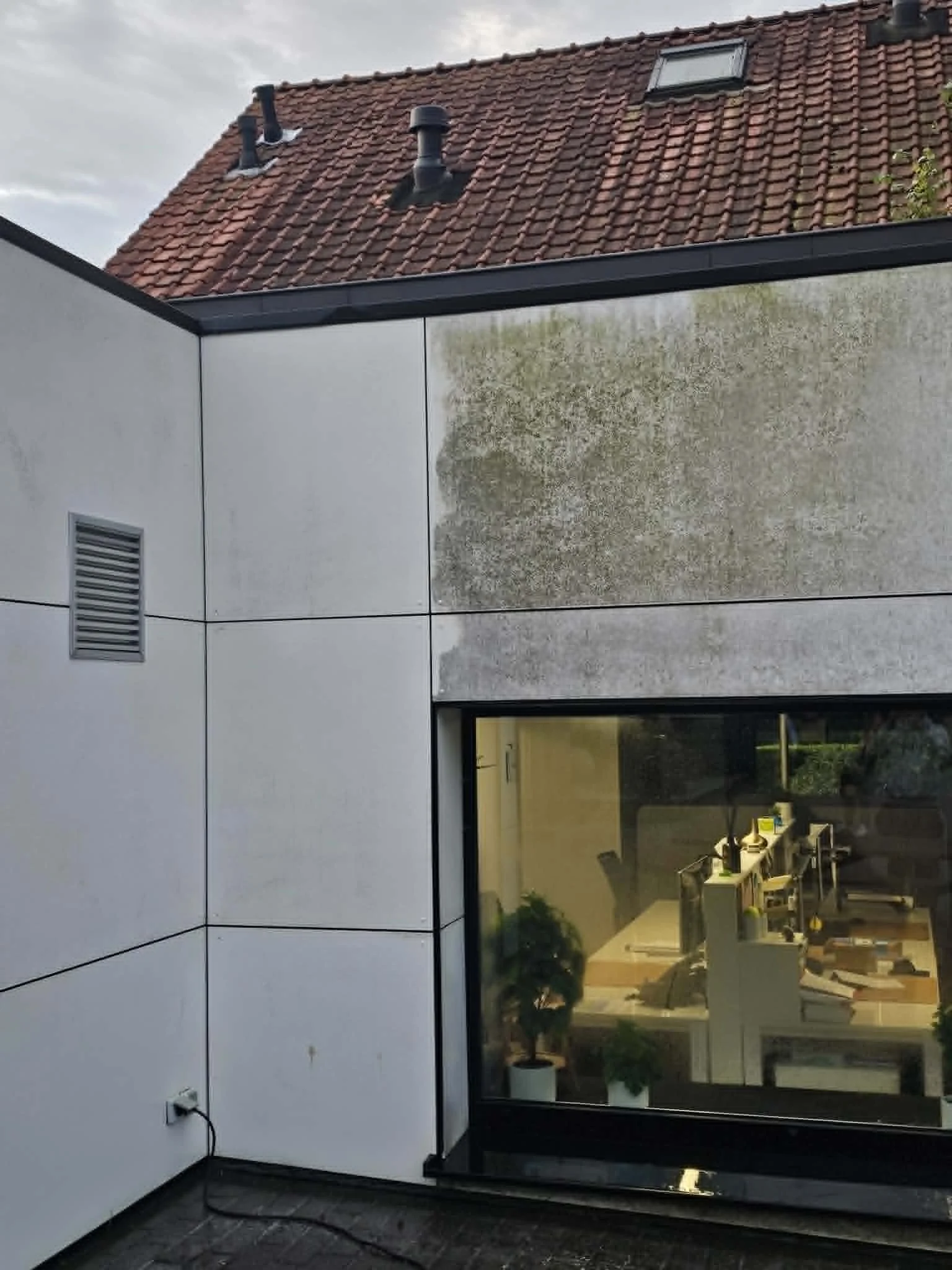 Een moderne woning met grote glazen ramen en witte gevelpanelen, tuin met groen tapijt en meubels binnen zichtbaar door de ramen.