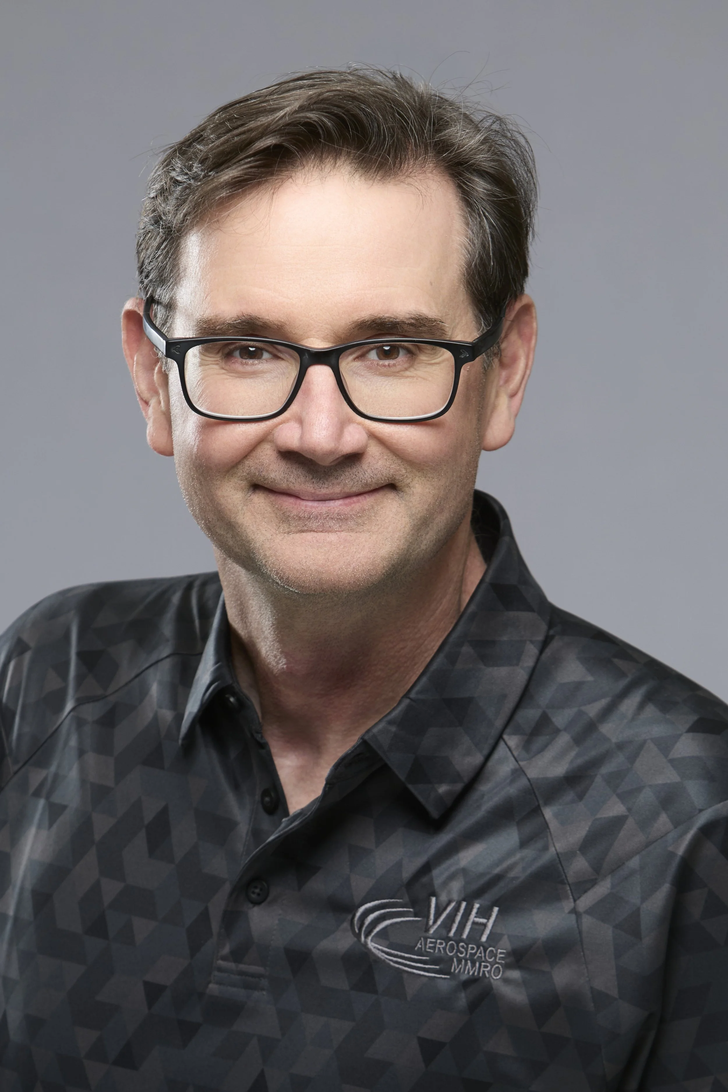 A man with short brown hair and a slight smile wearing a black collared shirt with a logo that says 'VIH Aerospace' on it, against a blurred background of warm lights.