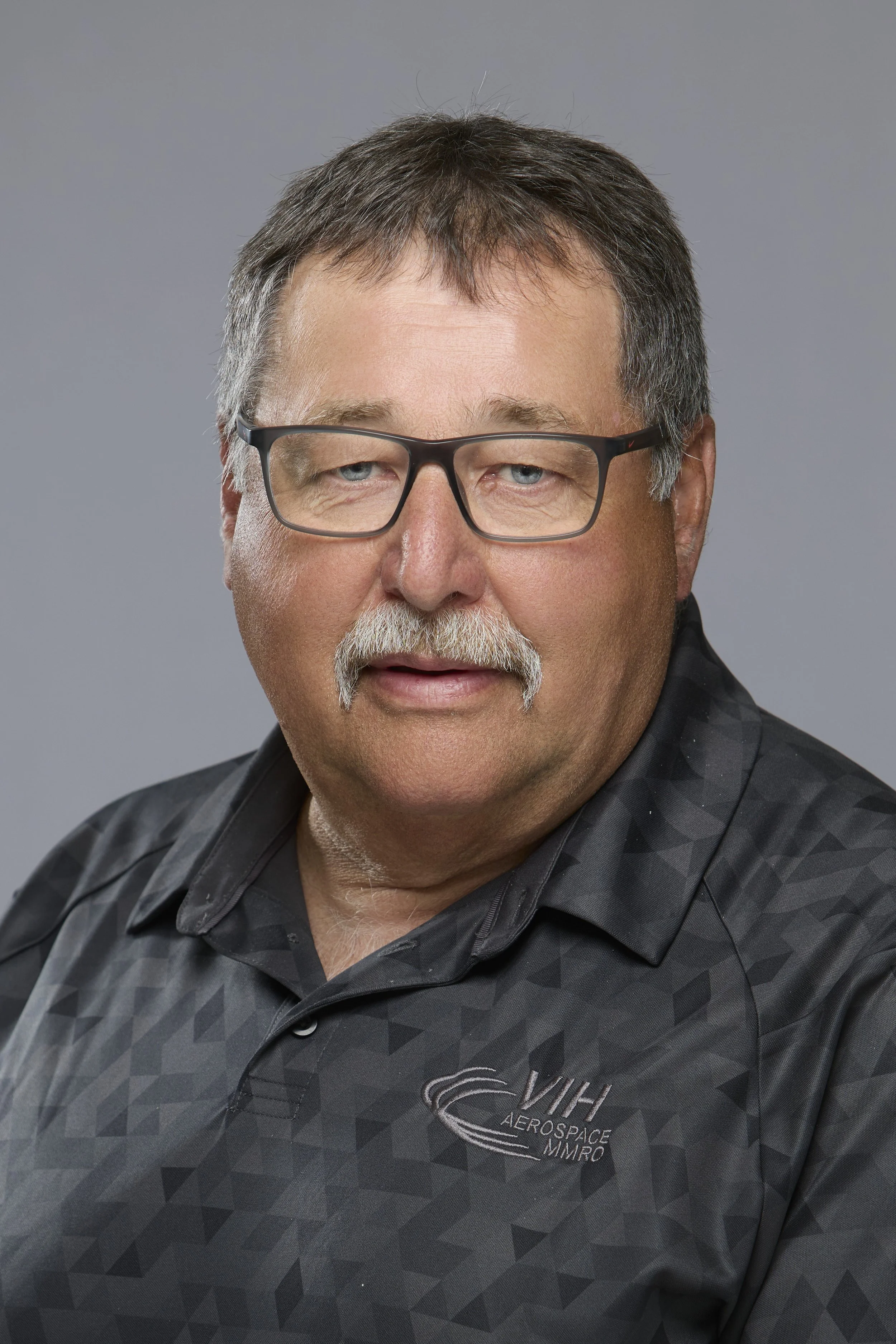 A middle-aged man with glasses, a mustache, and dark hair, smiling and wearing a black shirt with a VIH Aerospace logo, standing indoors with bokeh lights in the background.