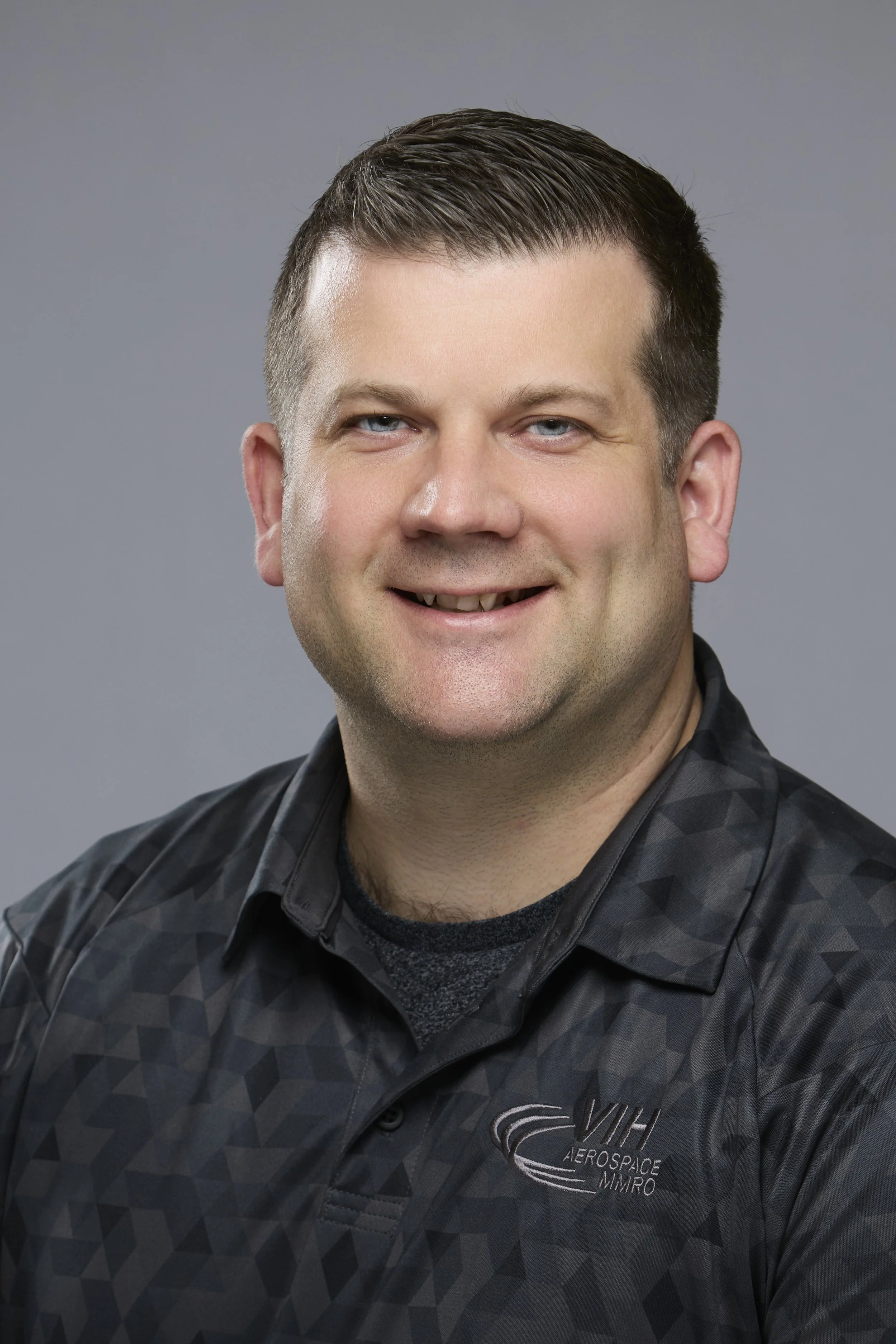 A man with short dark hair wearing a black polo shirt with the logo 'VIH Aerospace' on it, smiling at the camera, with a blurred background of warm lights.