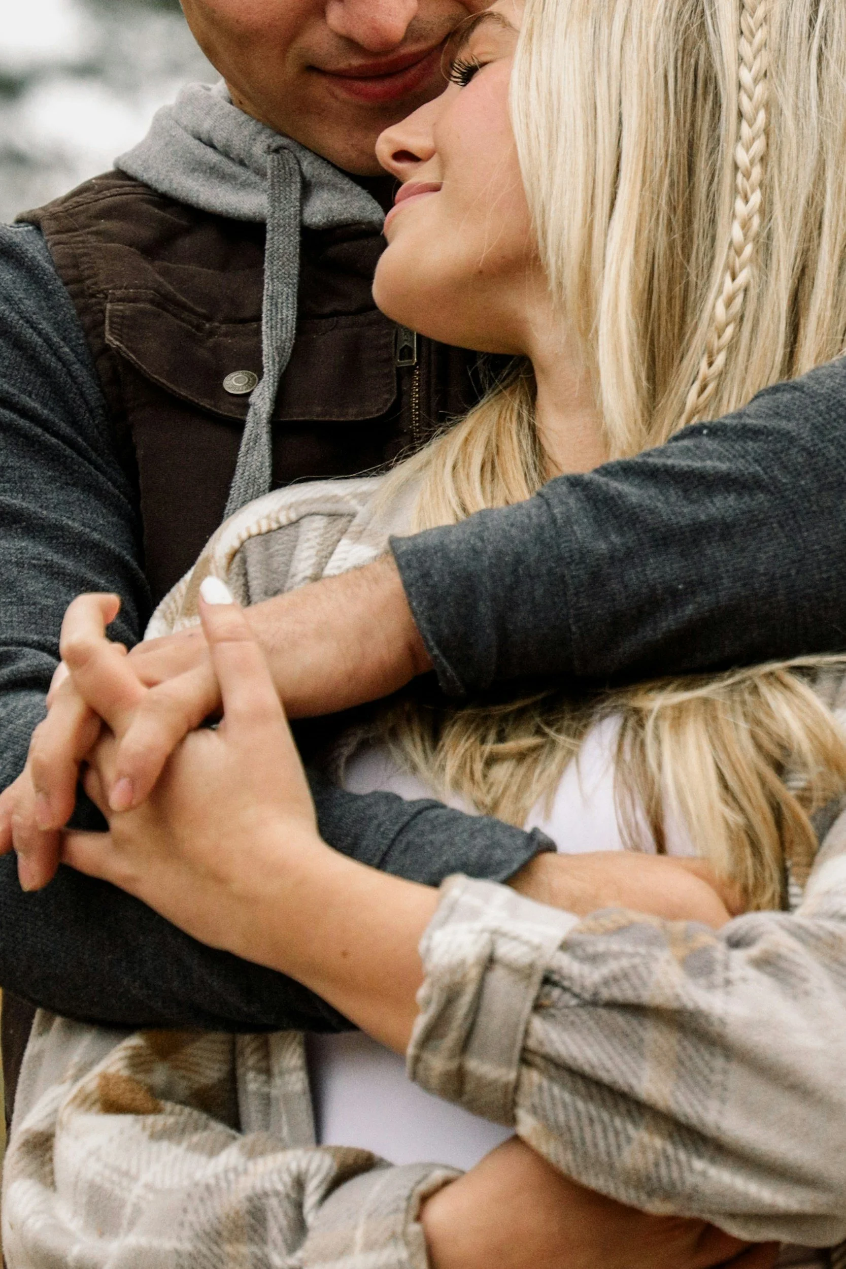 A young woman and a man embrace outdoors, with the woman smiling and resting her head on the man's chest. The man's face is partially visible, and they are holding hands.