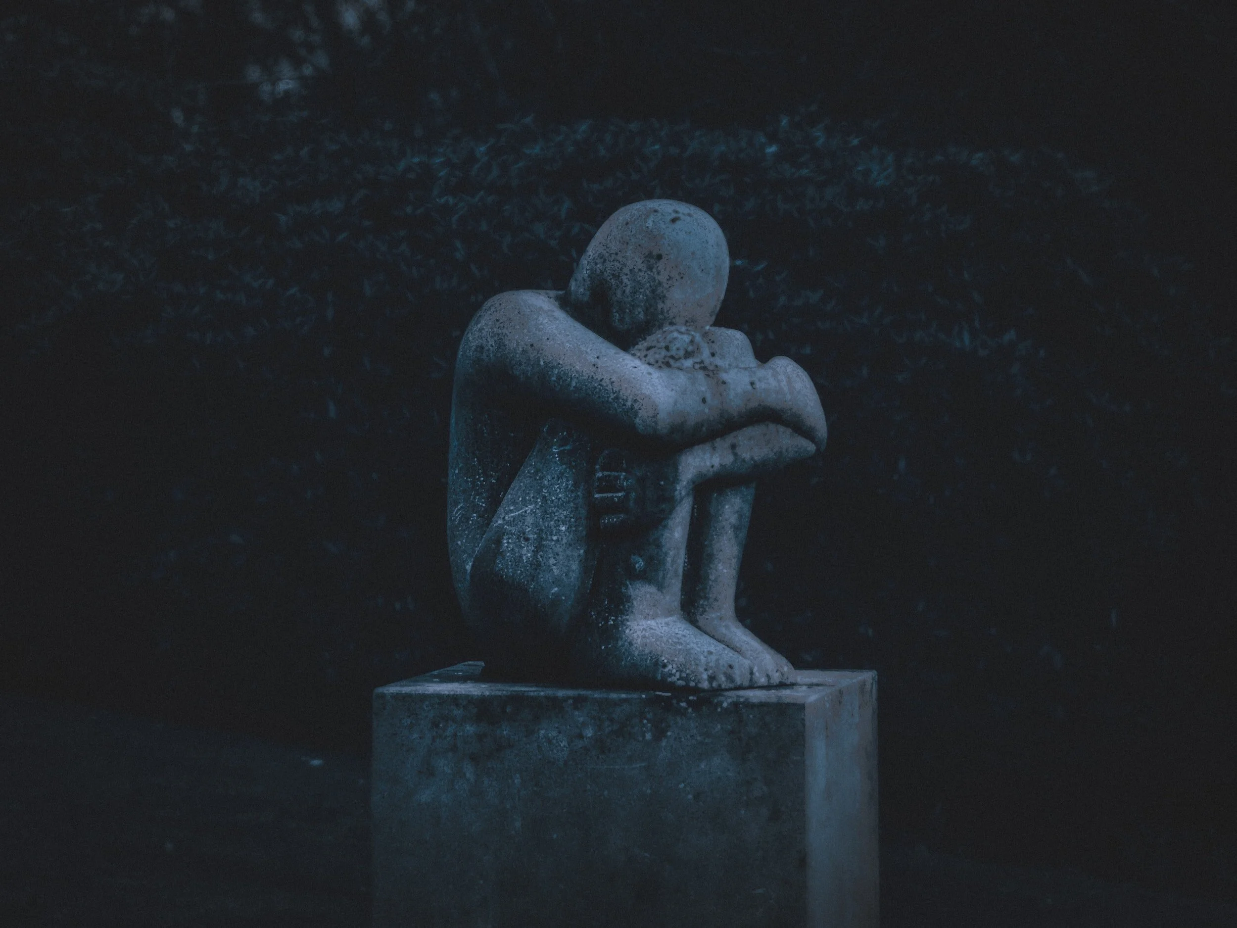A dark, somber sculpture of a human figure with a rounded head, sitting with knees drawn up and arms wrapped around the legs, resting on a rectangular pedestal.