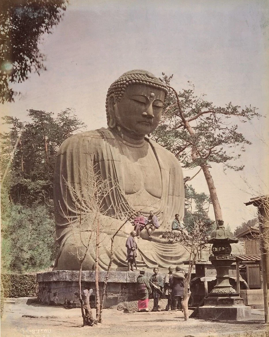 A large stone statue of Buddha, surrounded by a group of people, trees, and a small structure, in an outdoor setting.