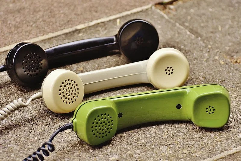 Three vintage rotary phones in black, cream, and green colors placed on a gravel path.