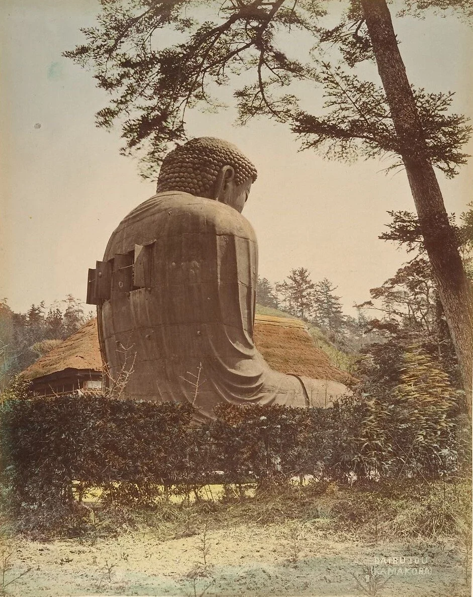 A large bronze Buddha statue sitting outdoors among trees and shrubs with a thatched-roof structure in the background.
