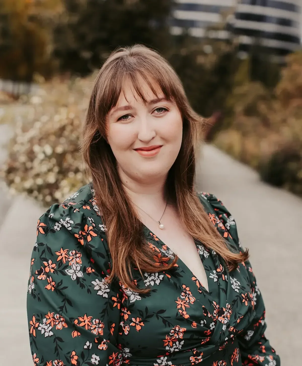 Kaitlyn Sheridan, a therapist with reddish-brown hair and bangs, wearing a dark floral dress and a pearl necklace, standing outdoors on a paved pathway with trees and buildings in the background.