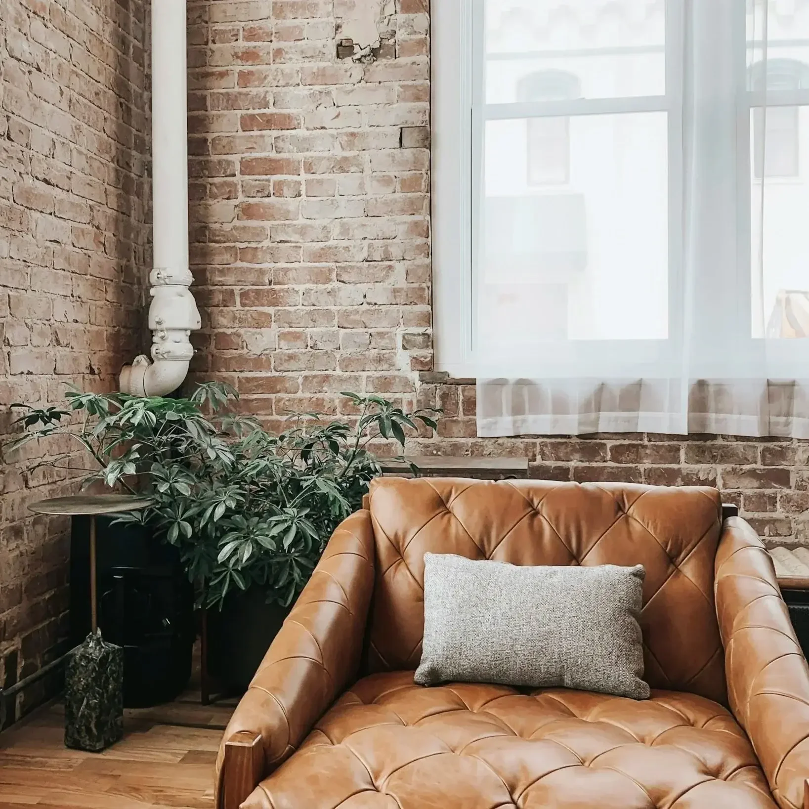 A cozy lounge area with a tan leather tufted sofa, a gray cushion, a potted plant, a side table with a marble base, and a large window with sheer white curtains unveiling a brick wall and building outside.