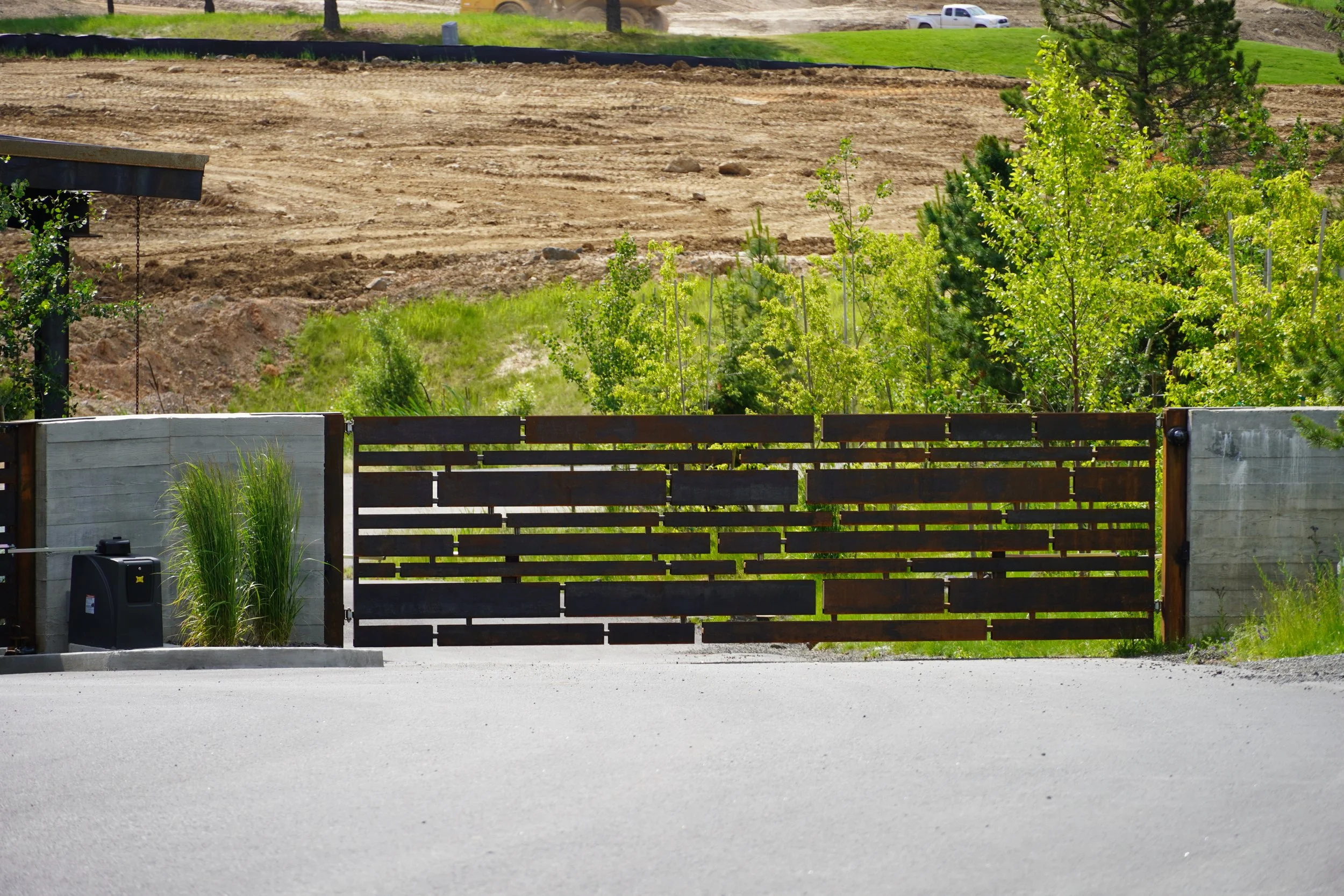 Black wooden gate with light gray concrete side panels, surrounded by green plants, and a dirt hillside in the background.