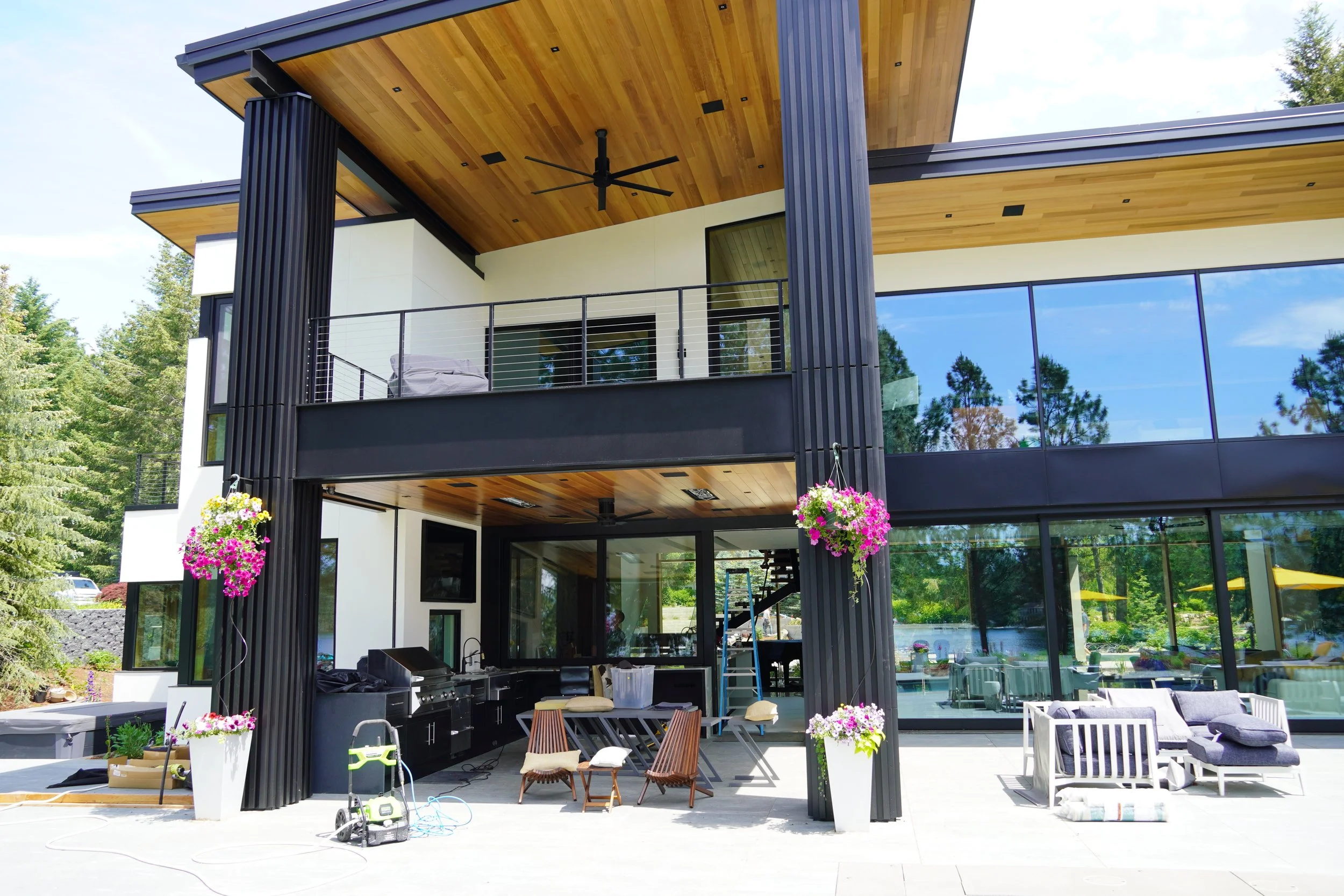 Modern multi-story house with large glass windows, black and white exterior, wooden ceiling, outdoor seating area, hanging flower baskets, and greenery surrounding the property.
