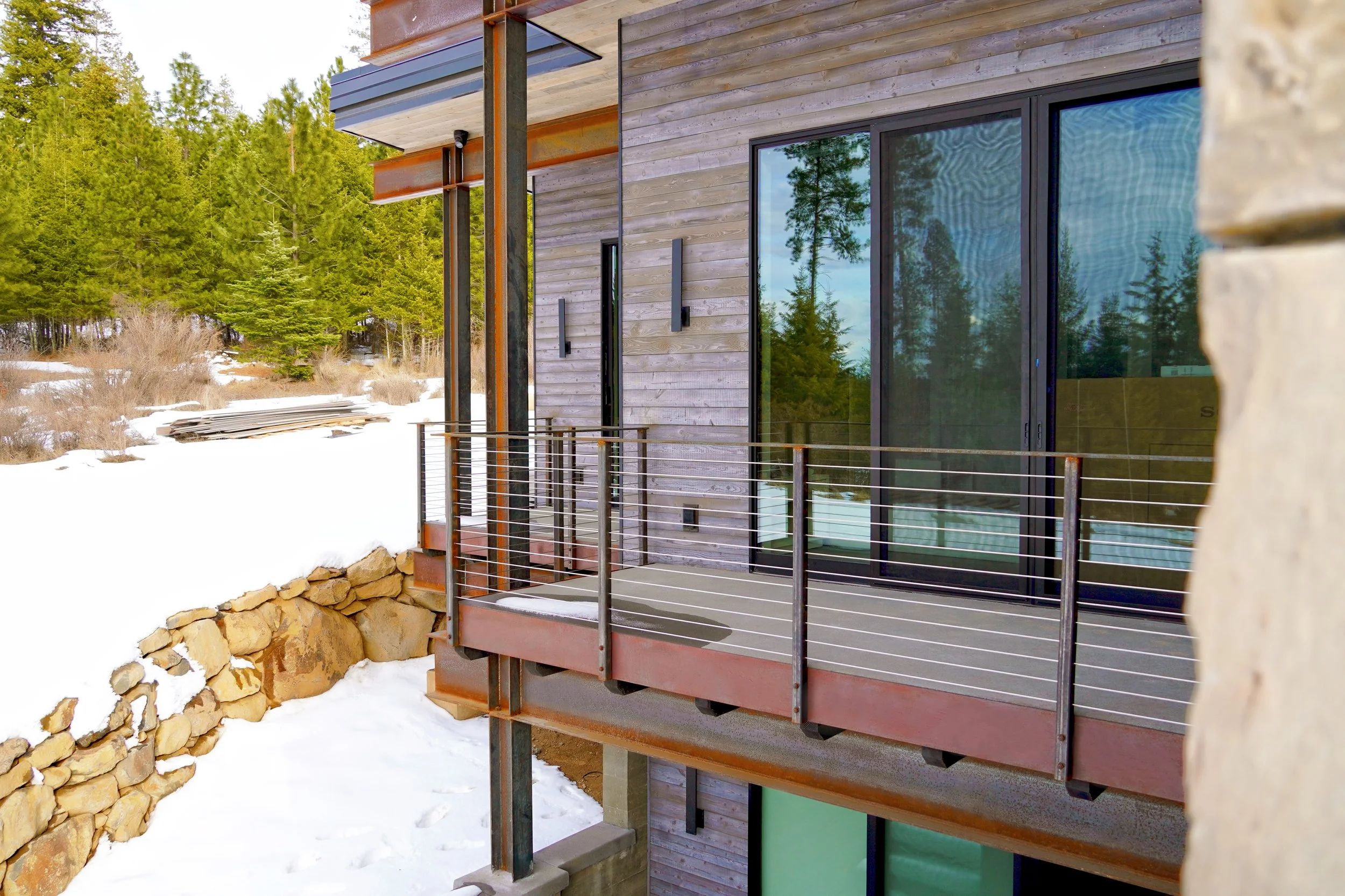 A modern balcony attached to a house with large glass sliding doors, a wood-paneled exterior, and metal railing, overlooking a snowy landscape with evergreen trees.