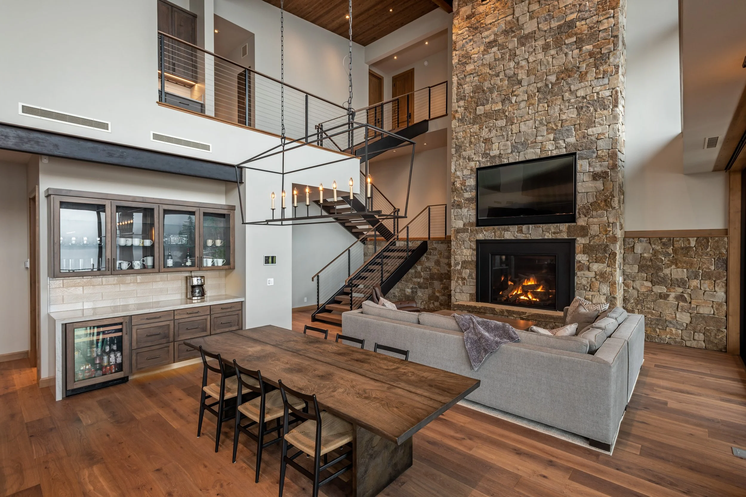 A spacious living room with hardwood floors, a large stone fireplace, a flat-screen TV above the fireplace, a gray sectional sofa, a wooden dining table with six chairs, a built-in cabinet with glass doors, a modern rectangular chandelier, and a stai