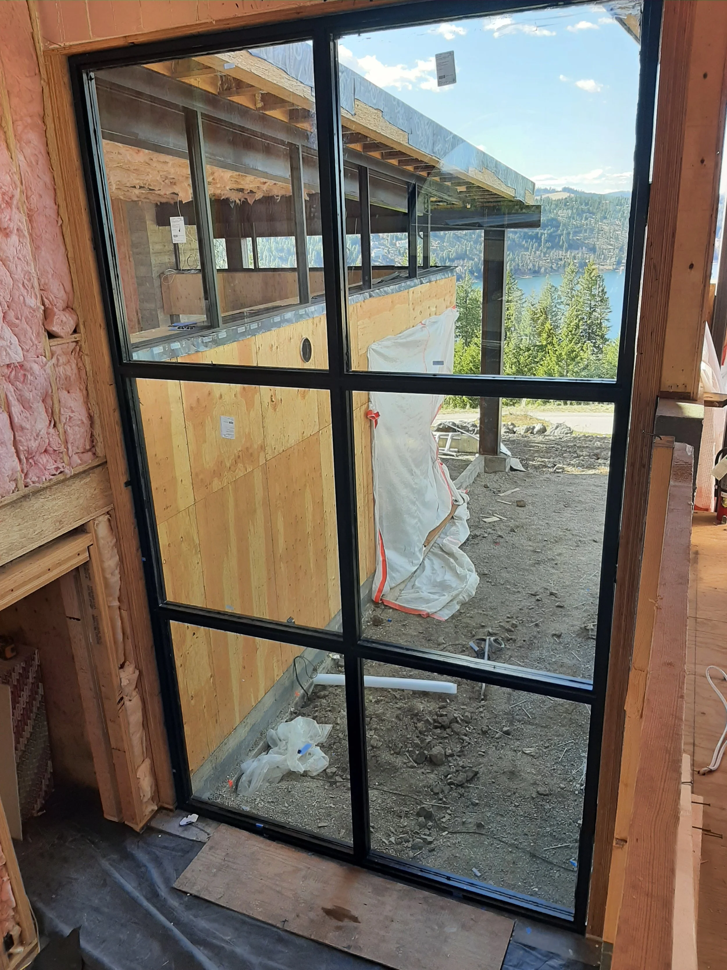 Interior view of a house under construction showing a black-framed glass door opening to an outdoor area with dirt and construction debris, with a scenic mountain landscape and lake in the background.
