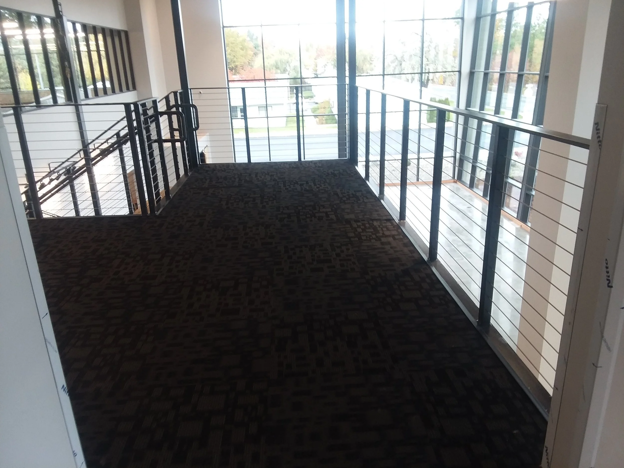 Interior view of a modern building with a balcony or mezzanine level, featuring a patterned dark carpet, metal railings, and large glass windows overlooking a suburban street.