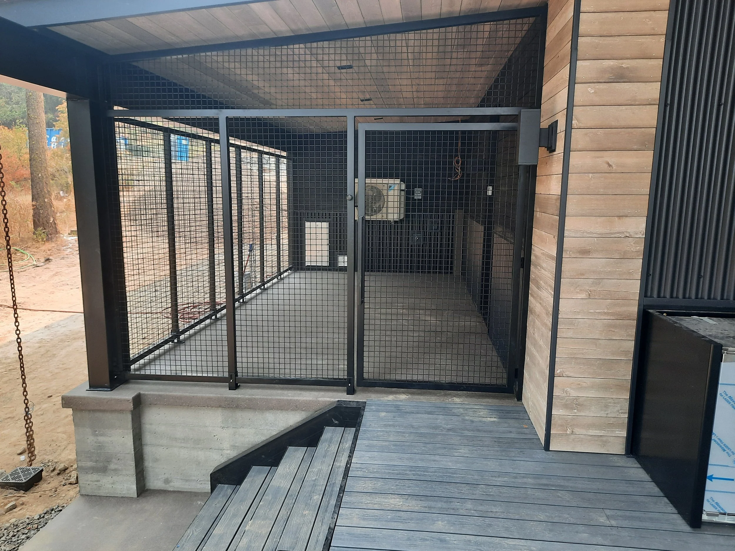 Outdoor dog kennel with black metal fencing, concrete base, wooden wall, and wooden deck, situated in a backyard with trees and a dirt area.