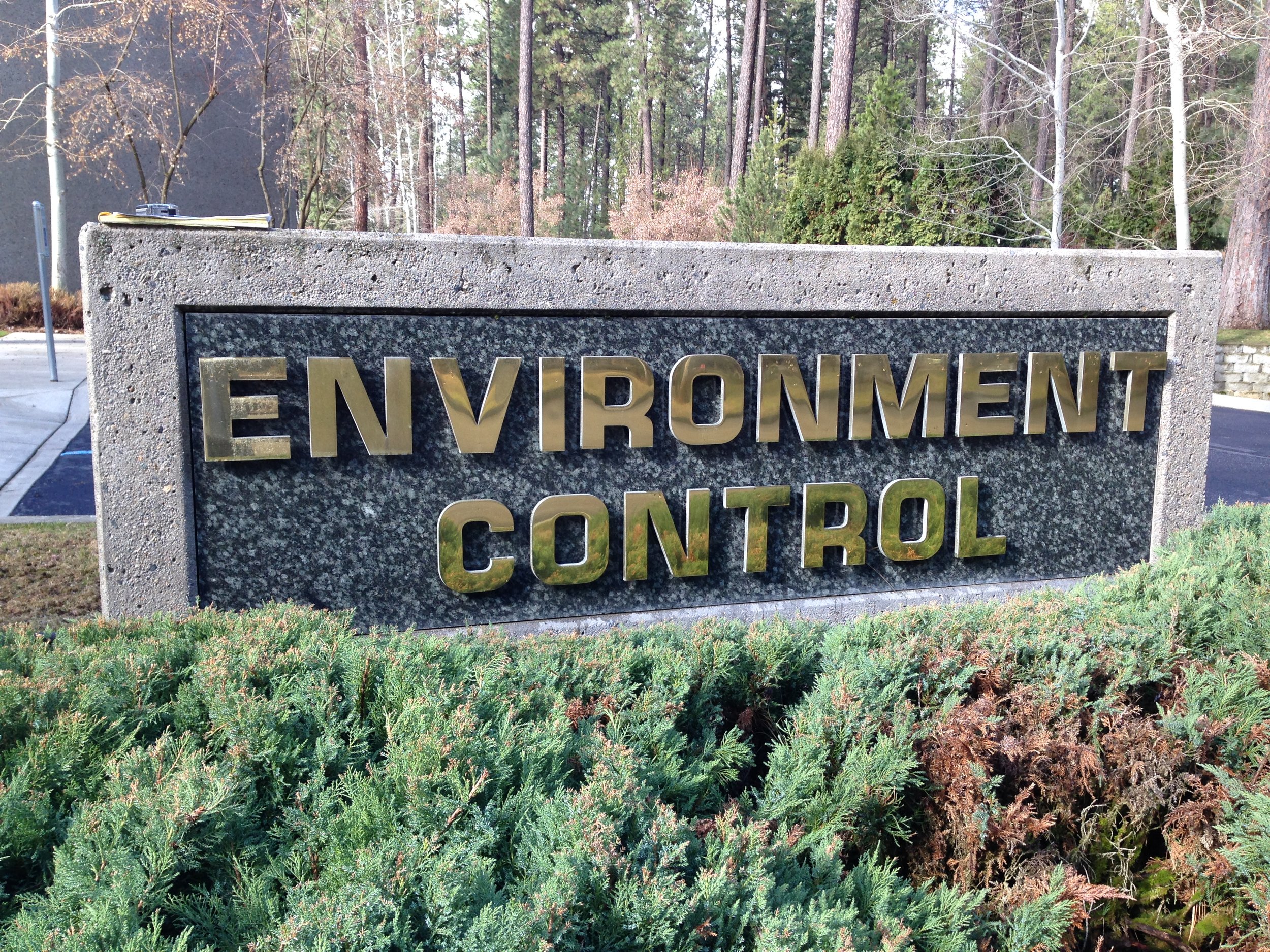 Sign that reads 'Environment Control' in gold letters on a granite background, situated in a landscaped area with bushes and trees in the background.