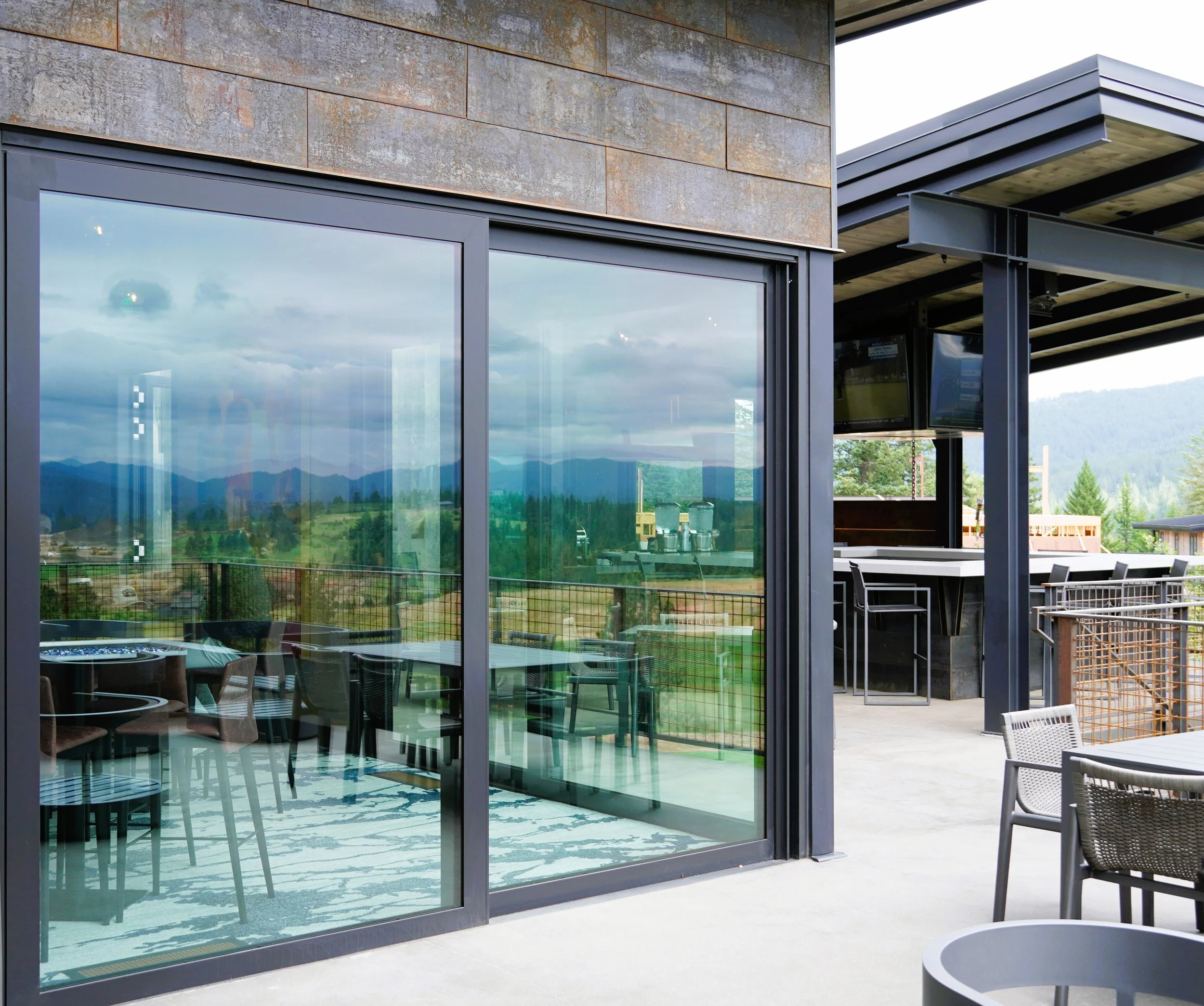 Modern outdoor patio with glass sliding doors reflecting scenic mountain view, with tables and chairs for dining or lounging.