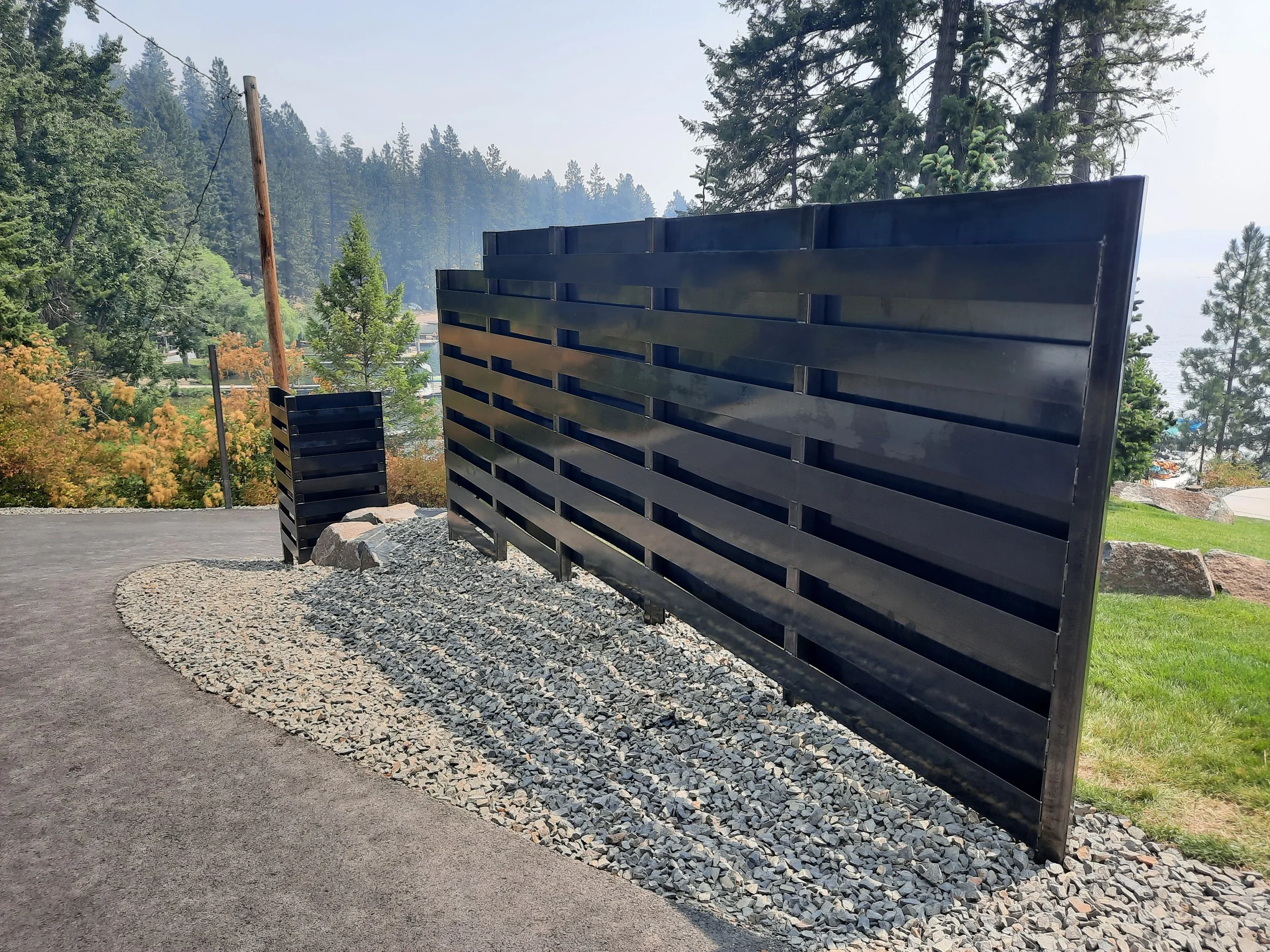 A black slatted privacy fence on a gravel base next to a paved pathway, with trees and a hillside in the background.