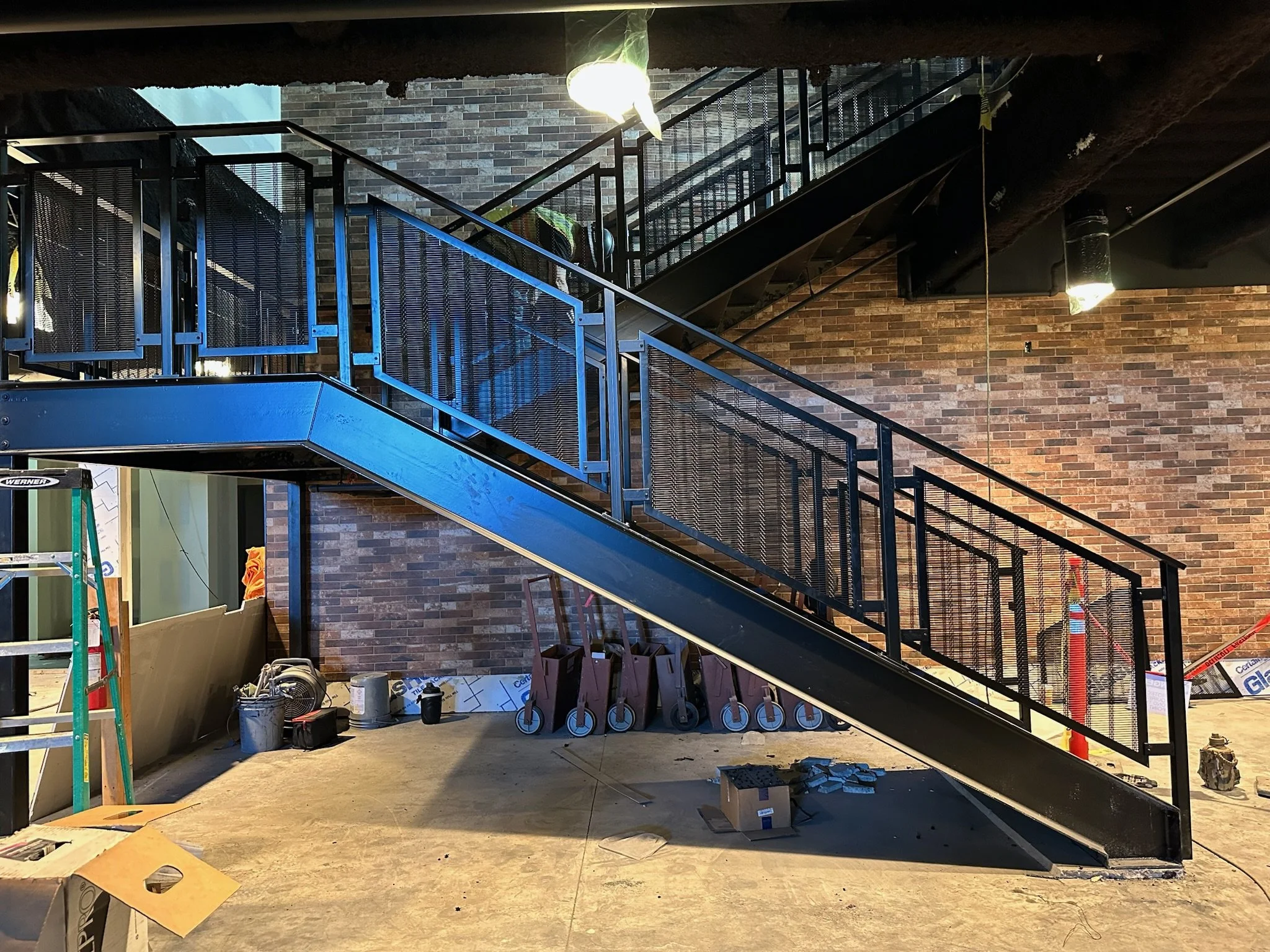 Construction site with black metal staircase and brick wall, construction tools and materials on the floor, and a ladder on the left side.