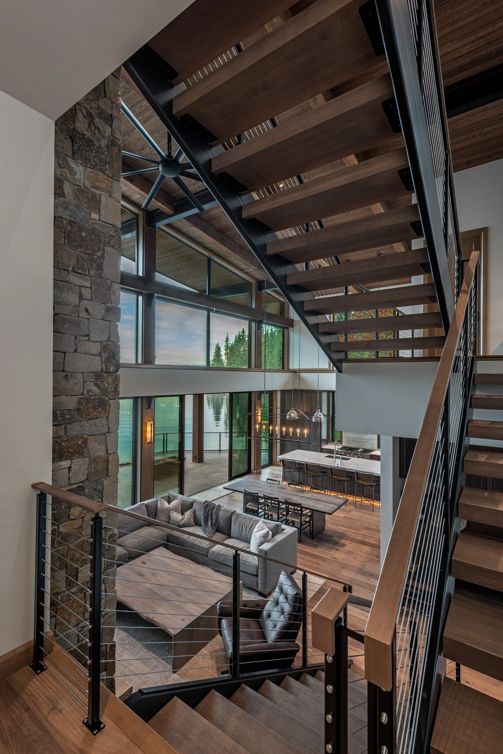 Interior view of a modern, open-concept living space with a staircase, large windows, a stone wall, a sofa, a dining table, and a kitchen with a waterfall island and bar stools.