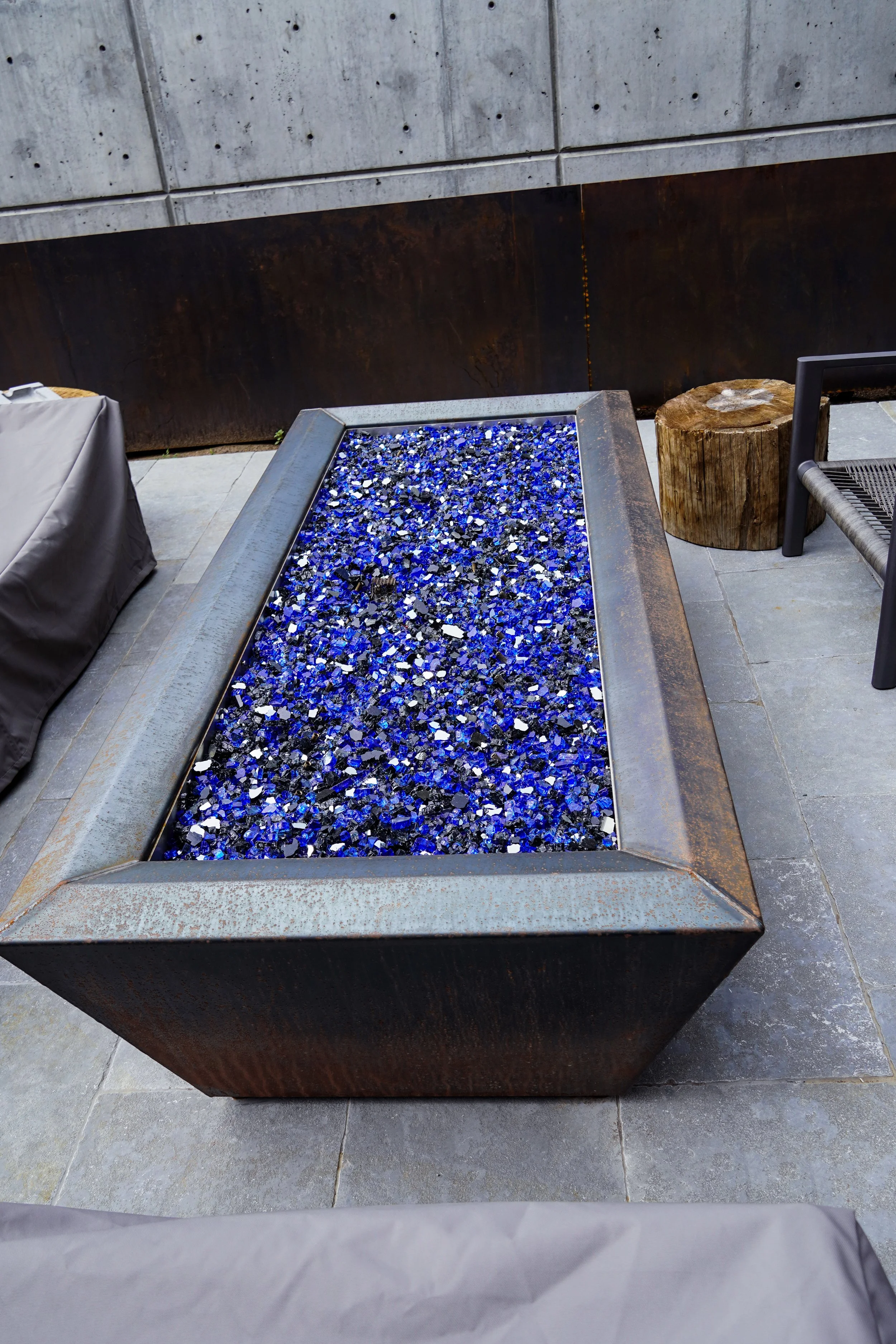 Outdoor fire pit with blue glass stones surrounded by stone patio, a tree stump, and outdoor furniture.