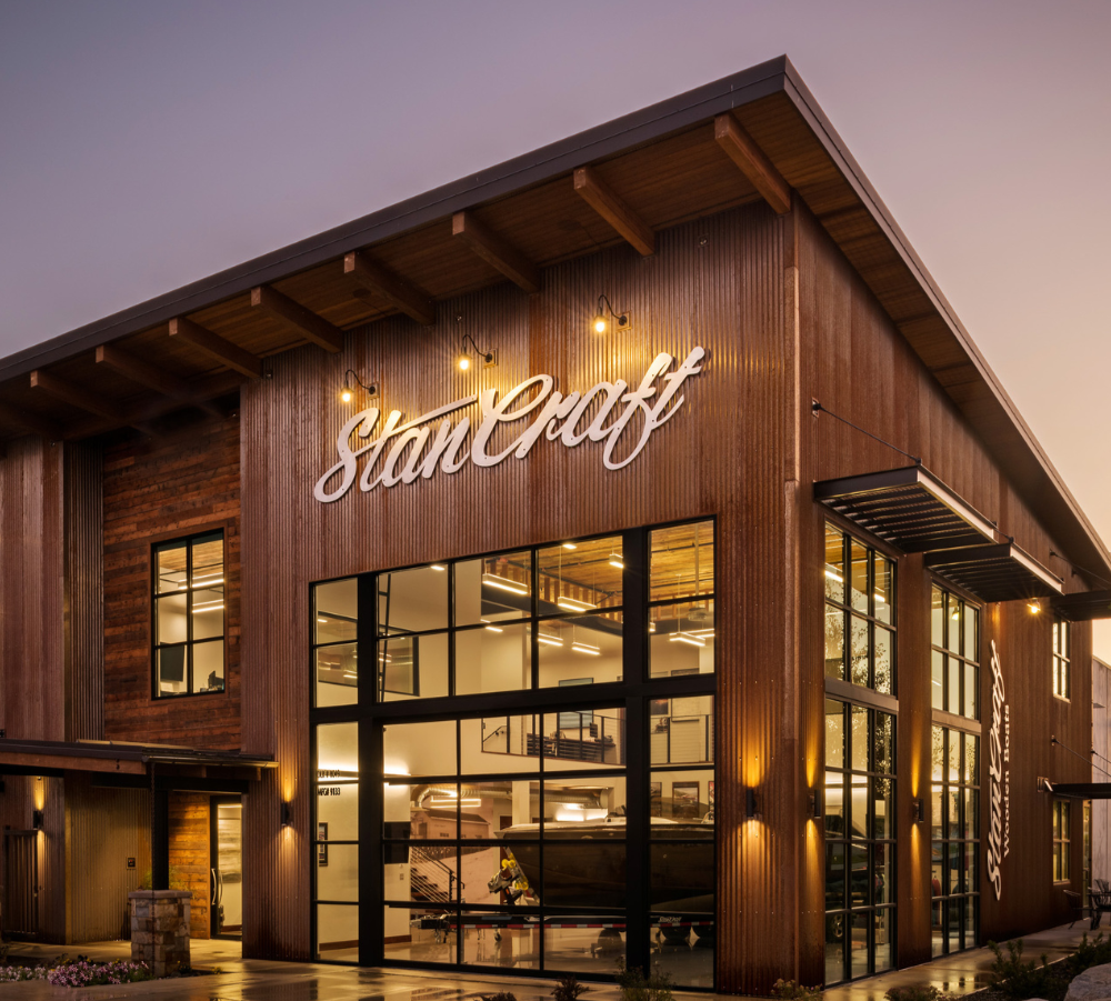 Exterior of a modern building with large glass windows and wooden siding, featuring the sign 'Stan Craft' in cursive lights, illuminated at dusk.