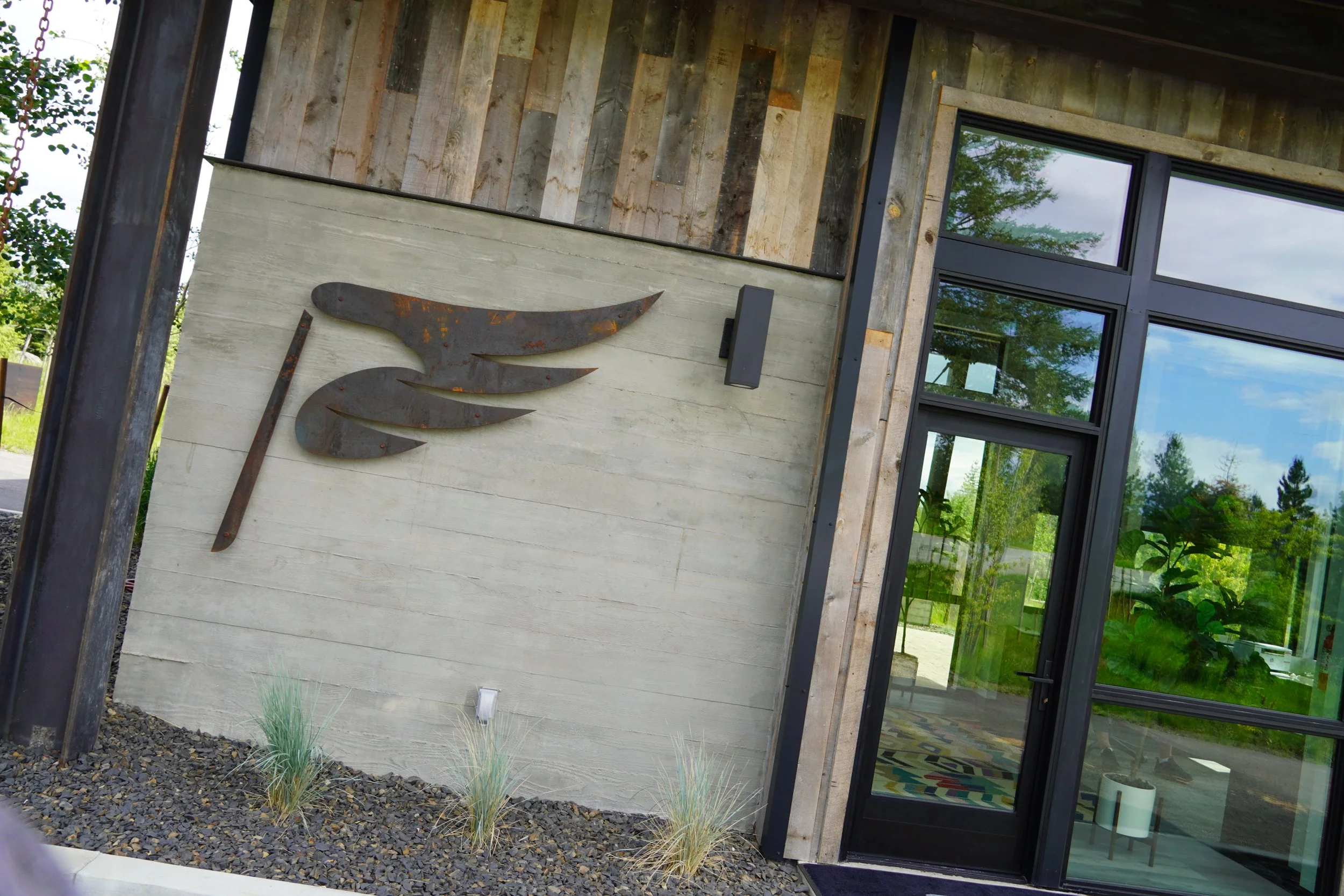 Exterior of a modern building with a metal wing-shaped sculpture on the wall. The wall is made of concrete and wood, with large glass windows reflecting trees and sky. Small plants are in front of the wall.