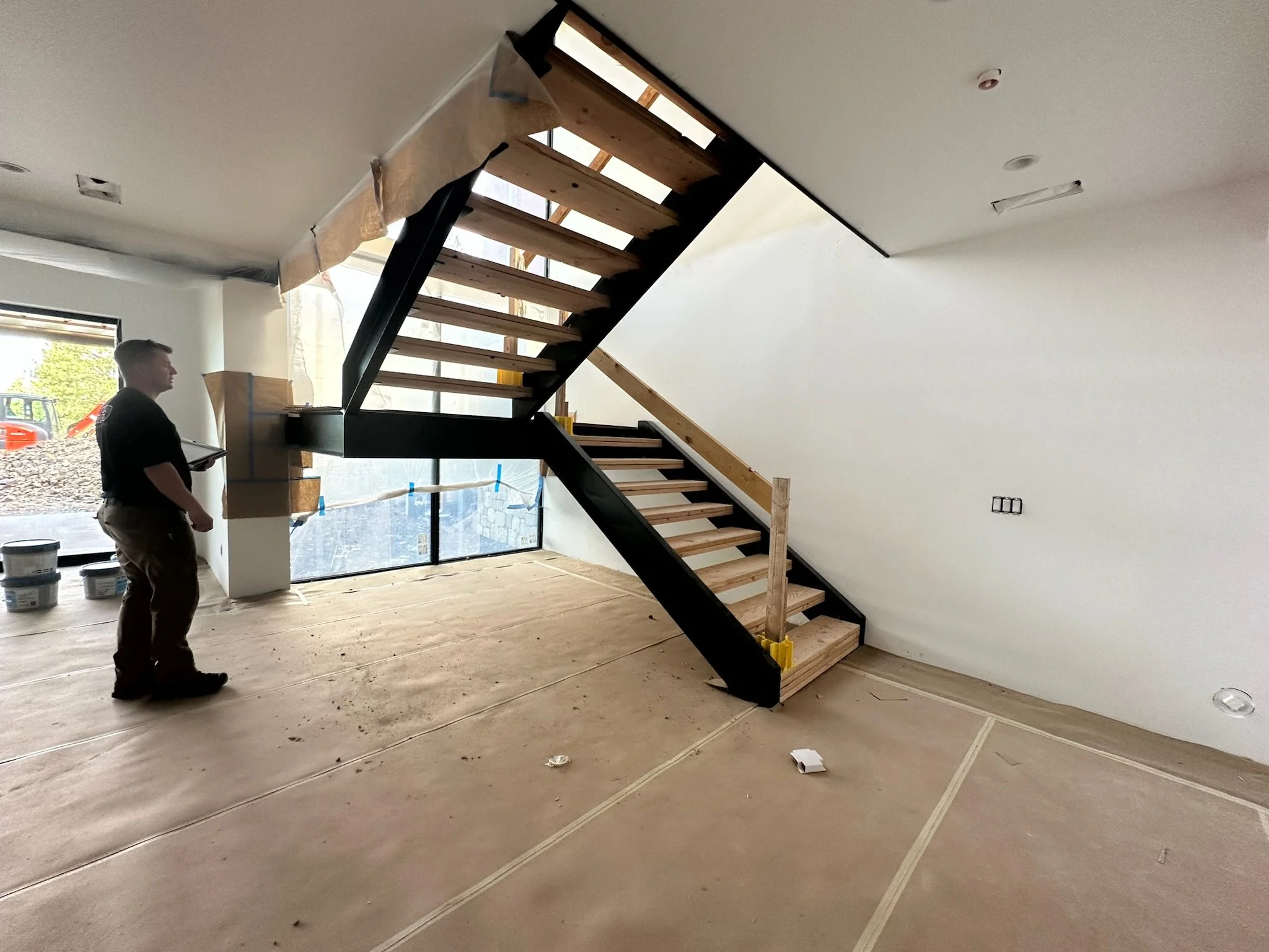 Interior of a house under construction showing a partially finished staircase with a person standing nearby holding a clipboard. The stairs are wooden with black framing, and construction materials and tools are visible in the space.