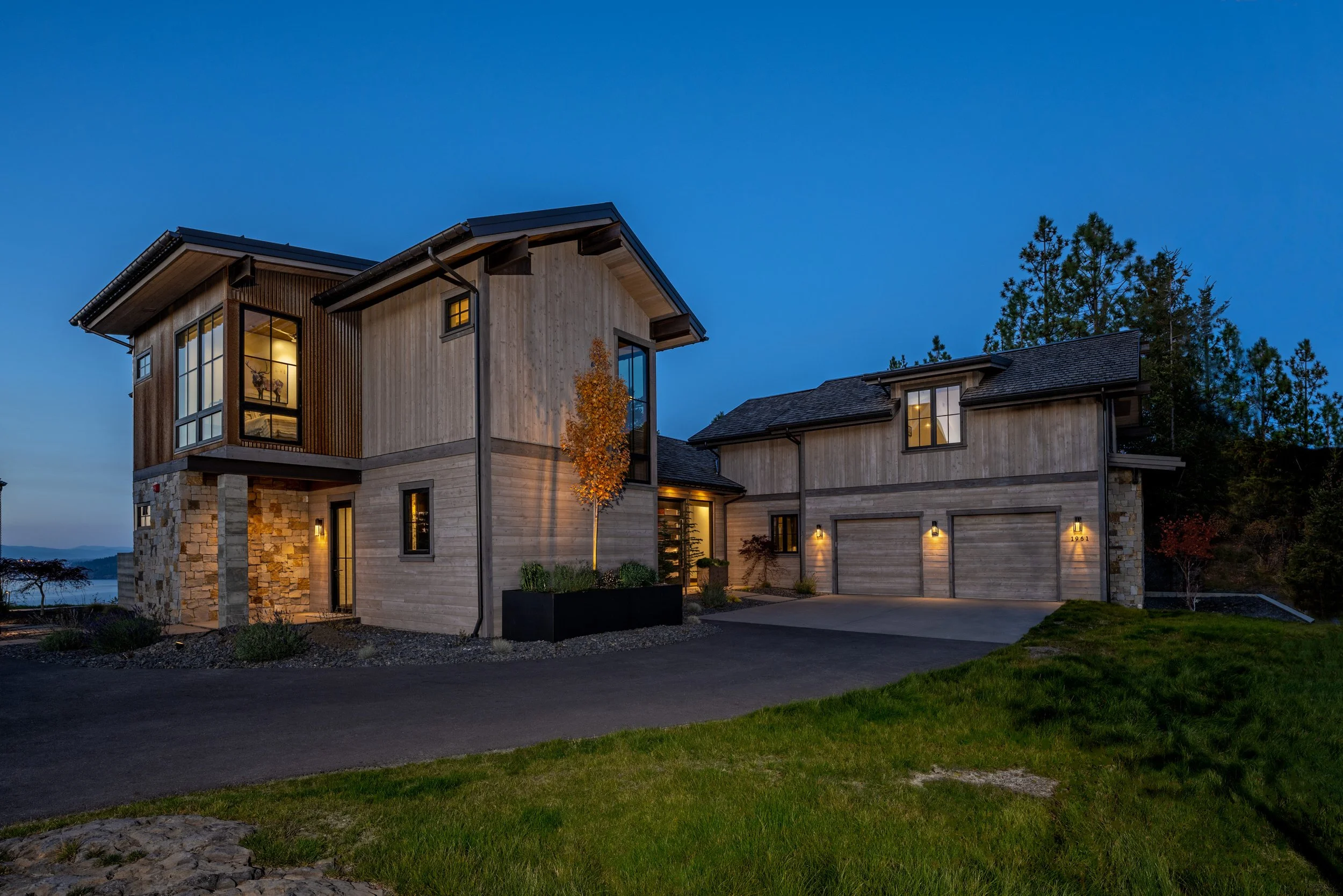 Modern two-story house with wooden siding, large windows, stone accents, and an attached garage at dusk.