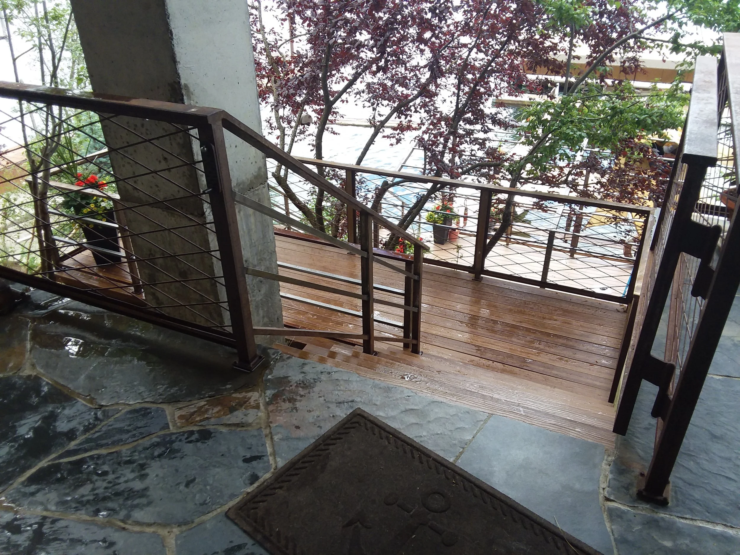 View of a wooden outdoor balcony with metal railings, surrounded by potted plants and trees, with a wet stone floor and a doormat in the foreground.