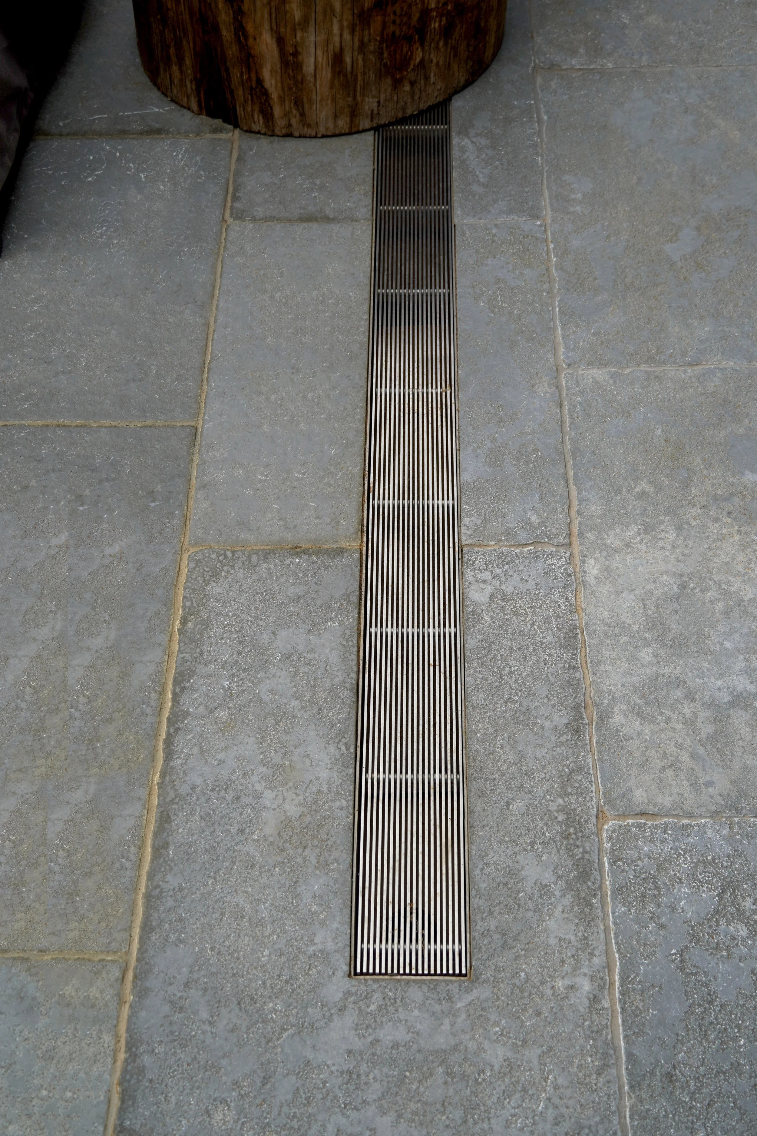 Close-up of a floor drain in a tiled floor, with gray stone tiles and a wooden object nearby.