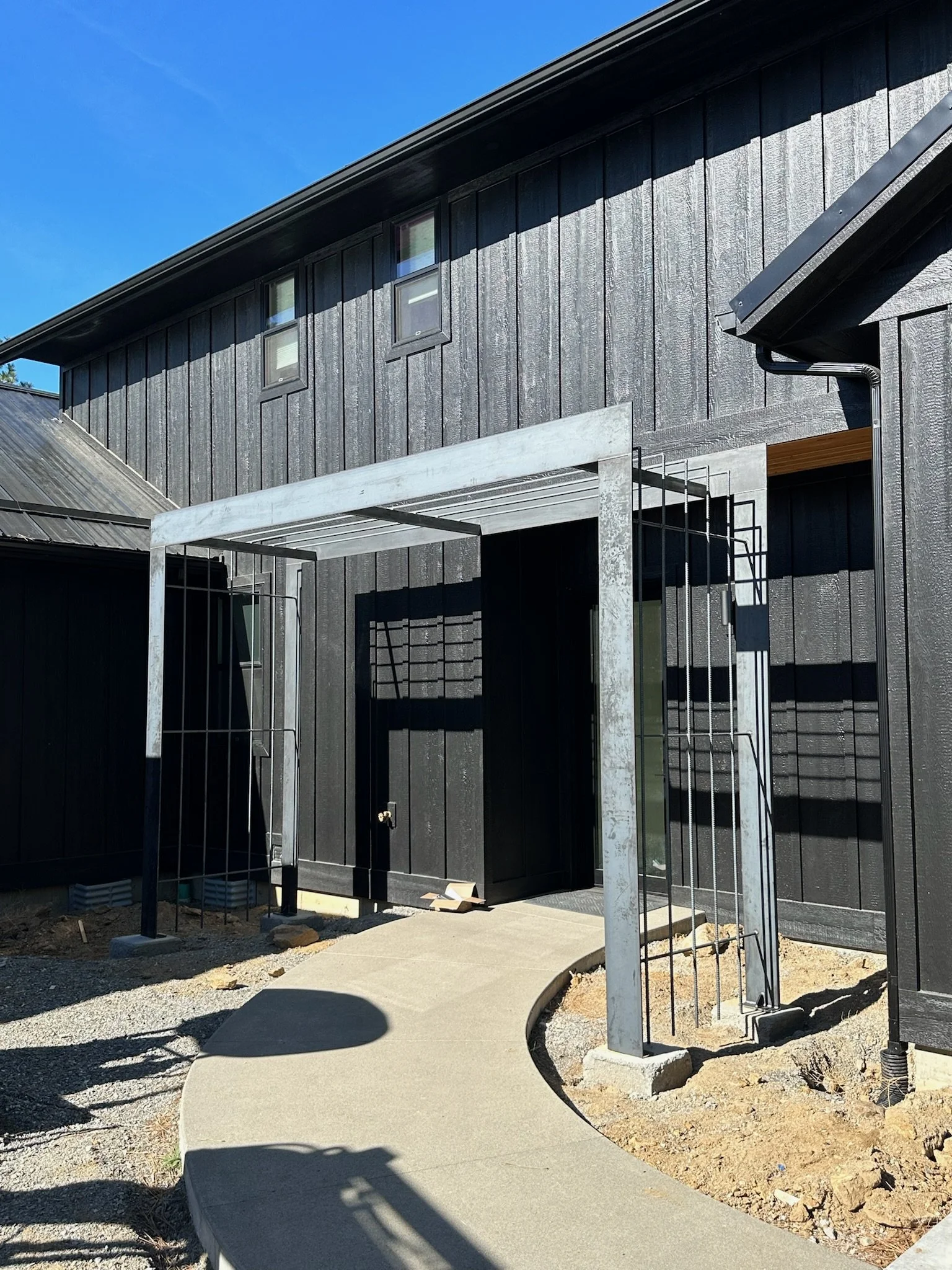 Newly constructed black wooden house with a concrete pathway leading to a black door, metal framework at the entrance, and a clear blue sky.