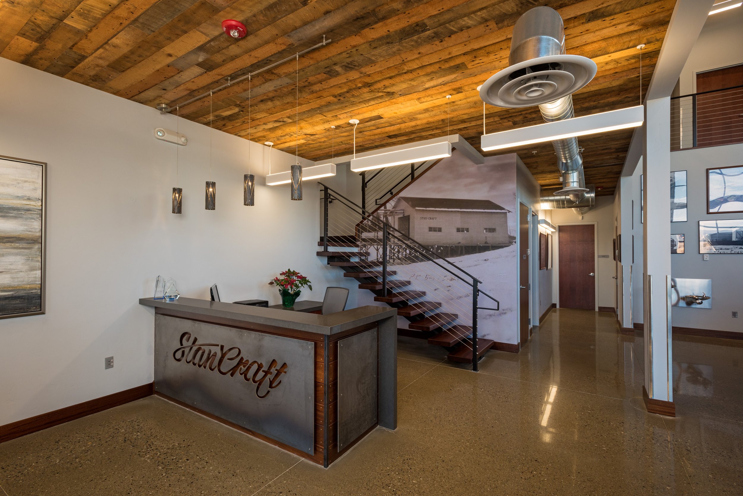 Modern reception area with a wooden ceiling, concrete floor, and white walls. There is a reception desk with the name 'StarCraft' on it, decorated with a small plant and glass awards. Behind the desk, a staircase with dark wooden steps and metal rail