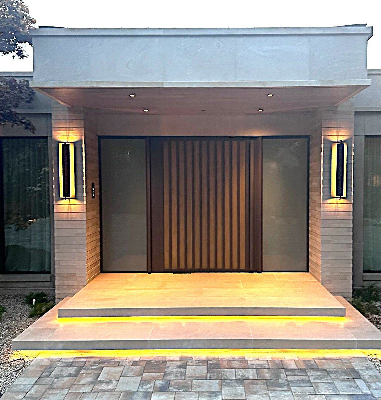 Modern house entrance with wooden door, stone steps, and two vertical wall-mounted lights, illuminated with yellow lighting.