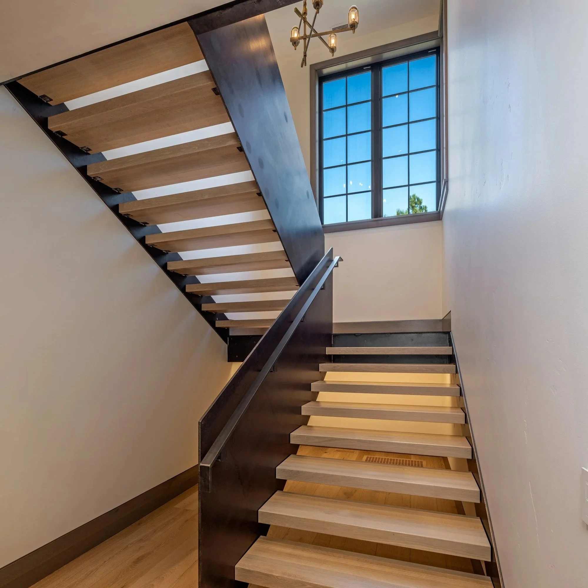 Modern staircase with wooden steps, black metal sides, and a wooden handrail, adjacent to a large window with black framing, seen from below, with a contemporary chandelier hanging from the ceiling.