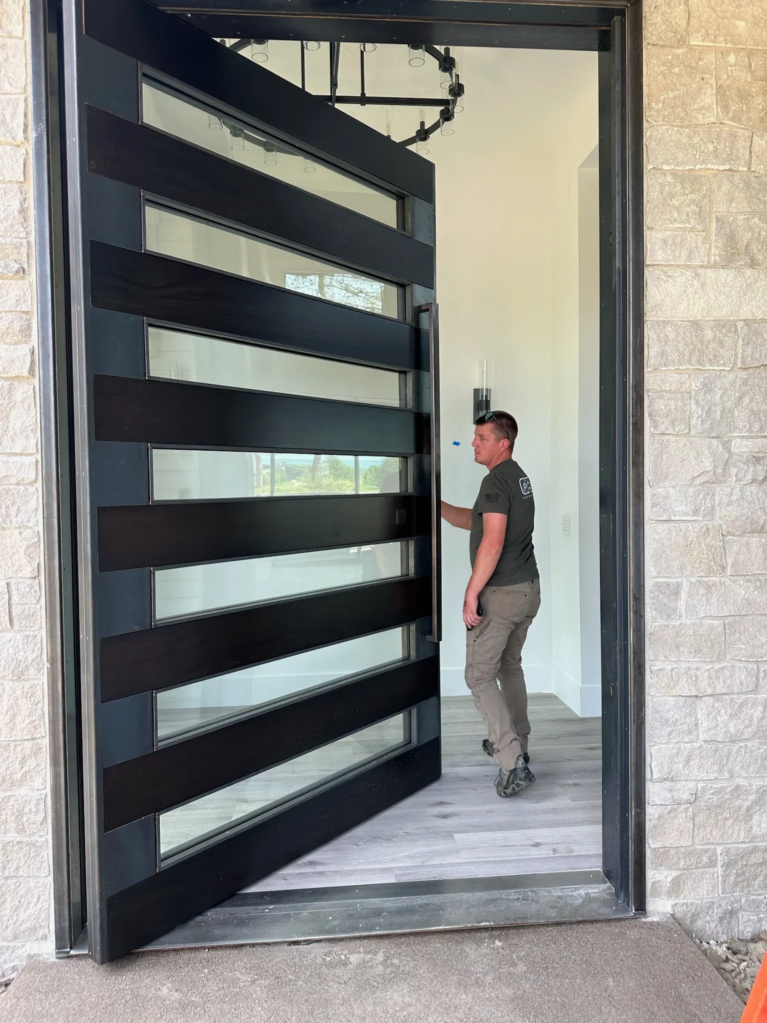 A man standing inside a modern home, near a large black front door with horizontal glass panels. The door is partially open, revealing a light-colored interior with a chandelier and large windows showing outdoor greenery.
