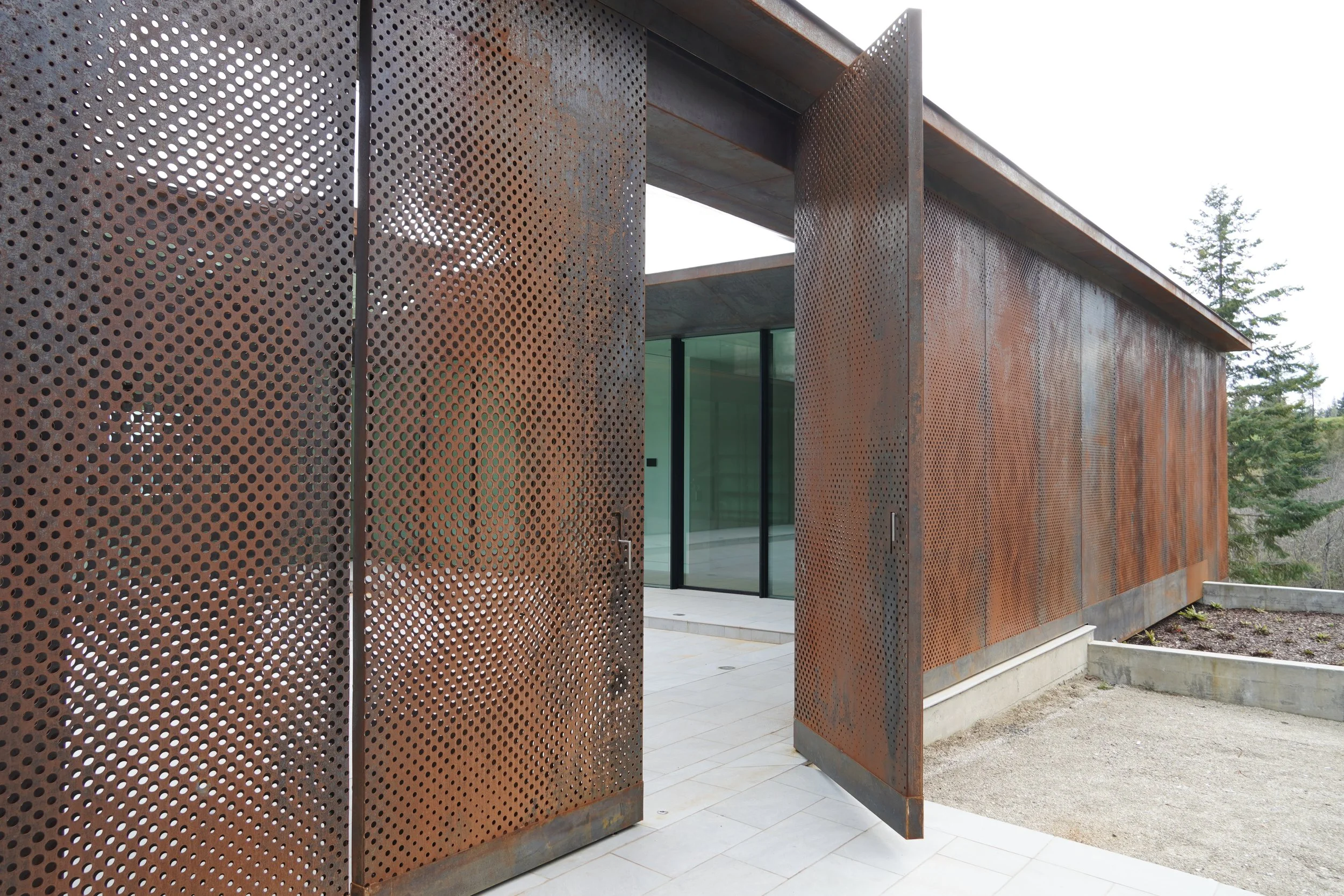 Modern building with rust-colored perforated metal walls and glass entrance, surrounded by trees.